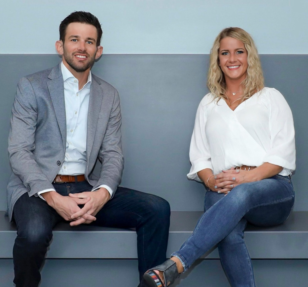 Young man with short dark hair and beard wearing a light gray blazer and white shirt, smiling and leaning on a railing indoors.