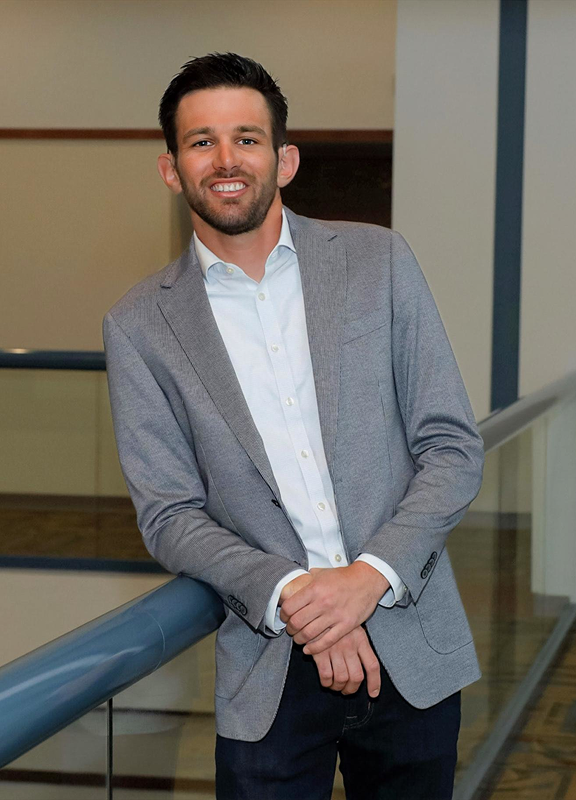 Young man smiling, leaning on a blue railing indoors, wearing a grey blazer, white shirt, and dark pants.