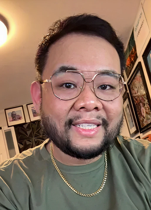 Smiling man with dark hair and beard wearing a gray blazer and white shirt leaning on a blue railing indoors.