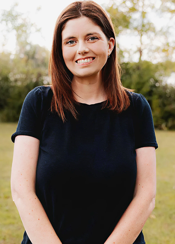 Smiling man with short dark hair and beard wearing a light gray blazer and white shirt, leaning on a blue railing indoors.