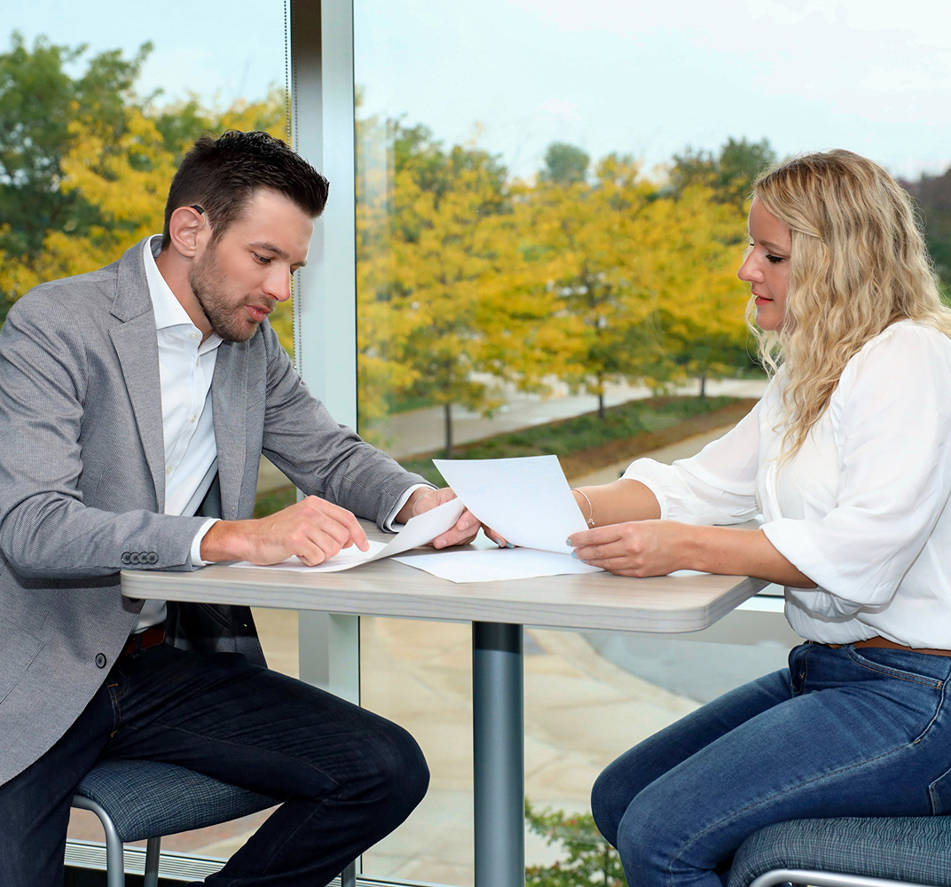 Smiling man in gray blazer and white shirt leaning on a railing indoors.
