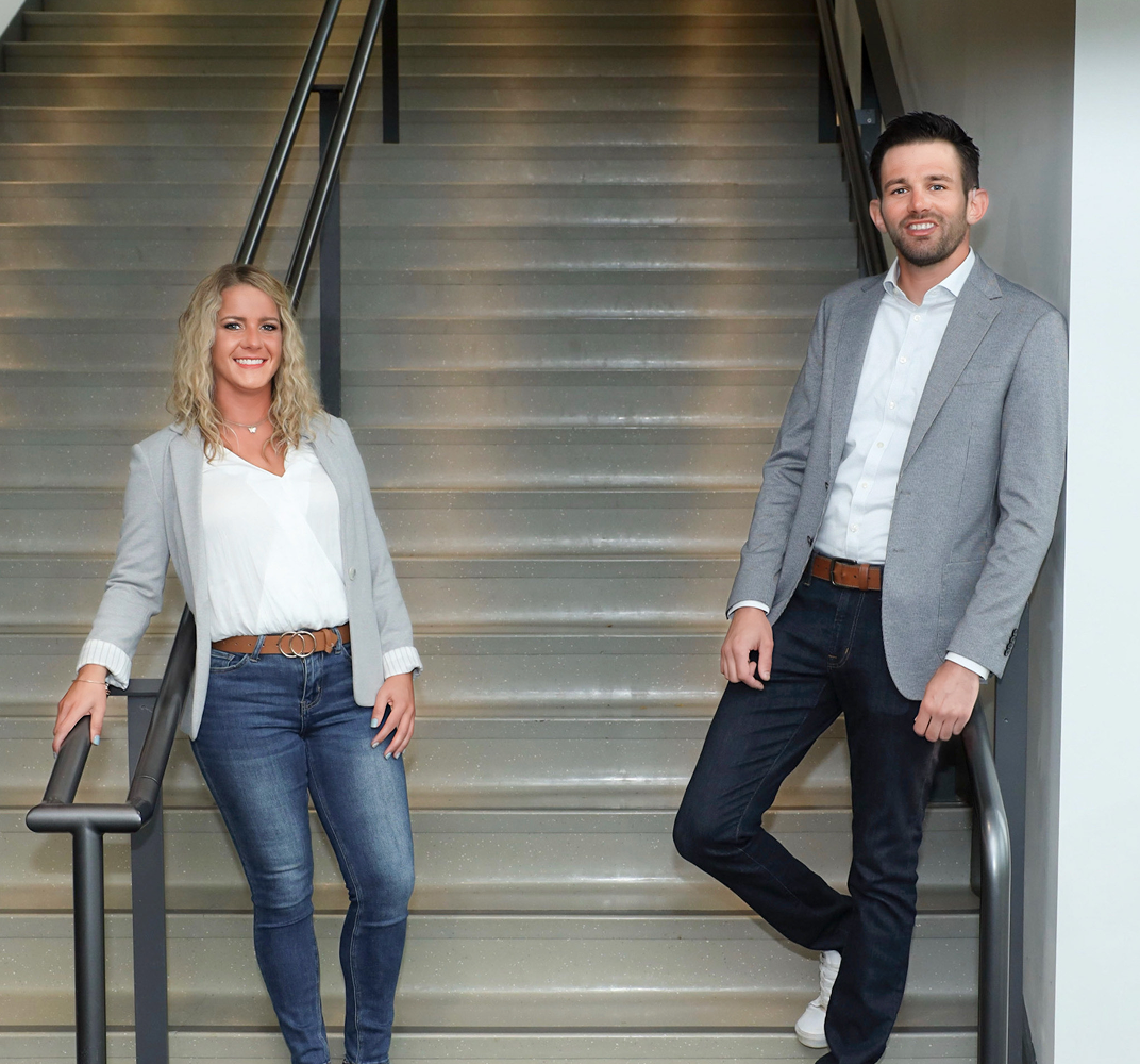Smiling man with dark hair and beard wearing a light gray blazer over a white shirt, leaning on a blue railing indoors.