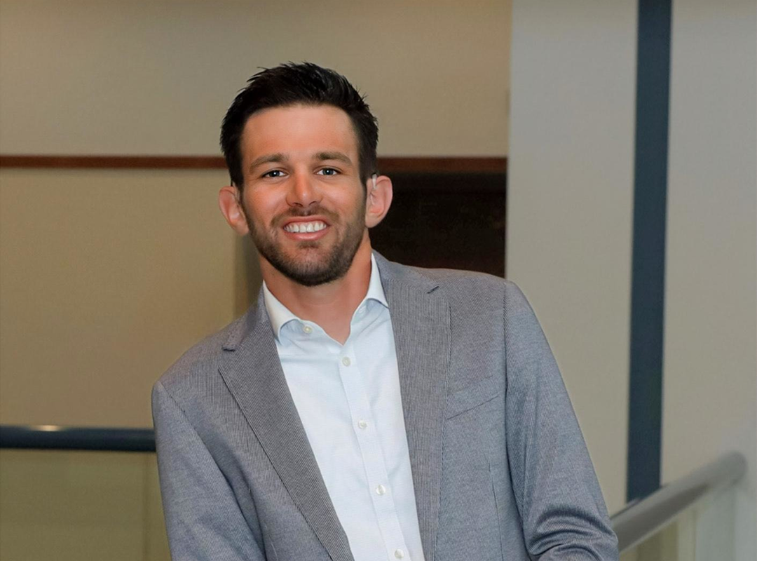 Smiling man wearing a gray blazer and white shirt, leaning on a railing indoors.