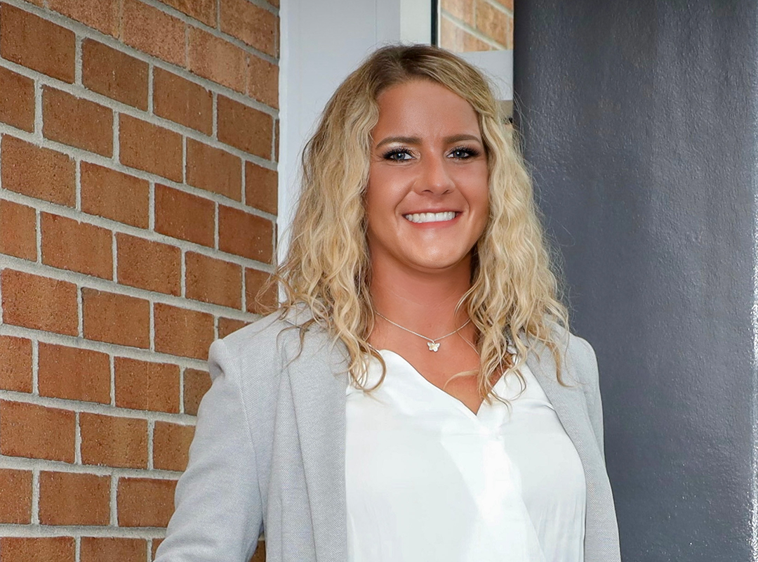 Smiling young man in a gray blazer and white shirt leaning on a blue railing indoors.