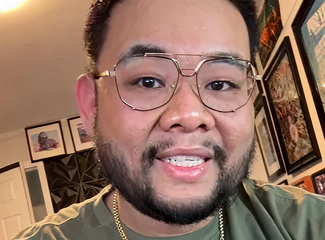 Smiling young man with short dark hair and beard wearing a gray blazer and white shirt, leaning on a railing indoors.
