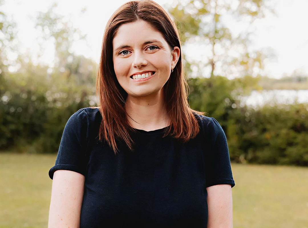 Smiling man with short dark hair and beard wearing a gray blazer and white shirt leaning on a blue railing indoors.