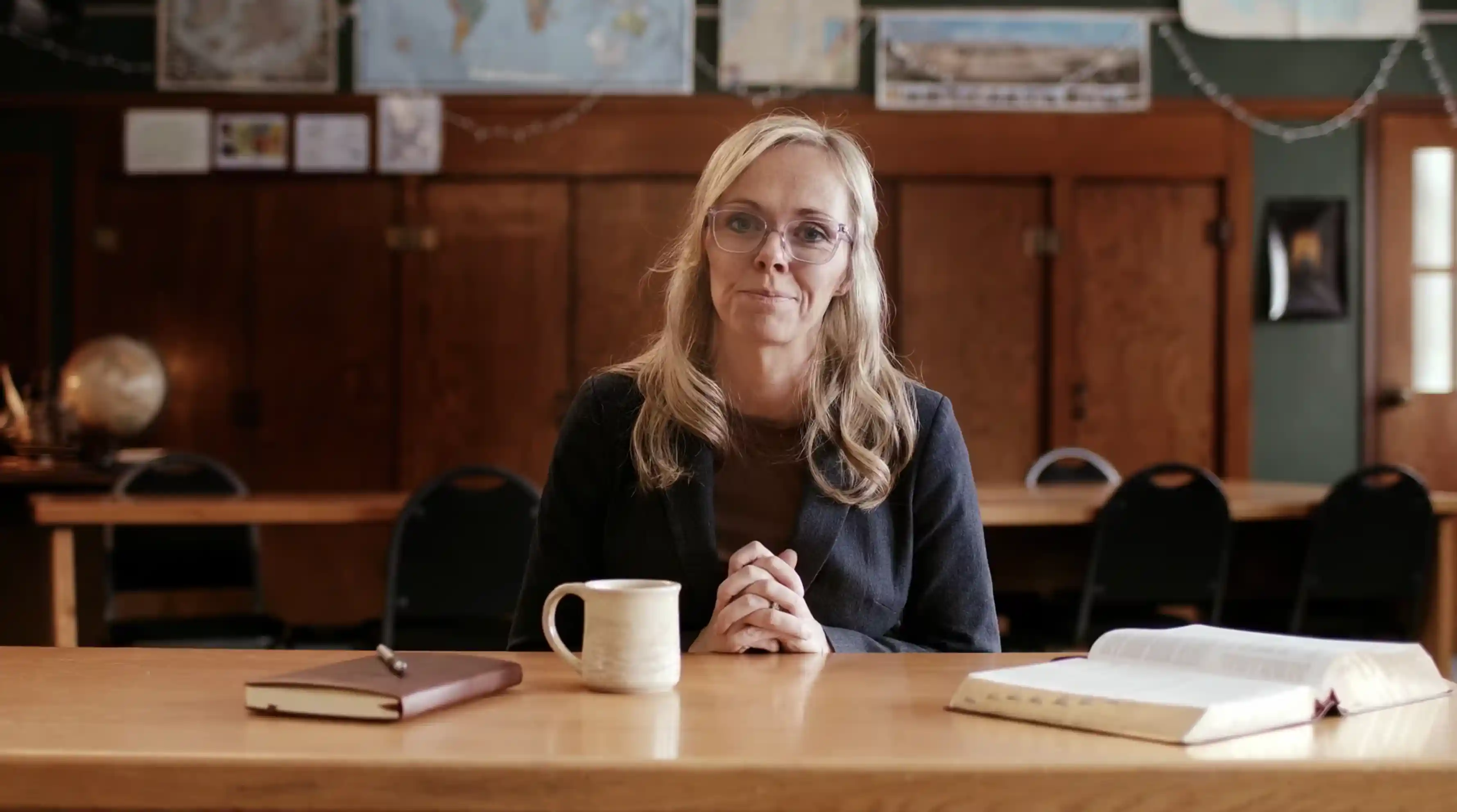 Blonde woman with glasses sitting at a desk with a mug, notebook, and open book in a classroom.