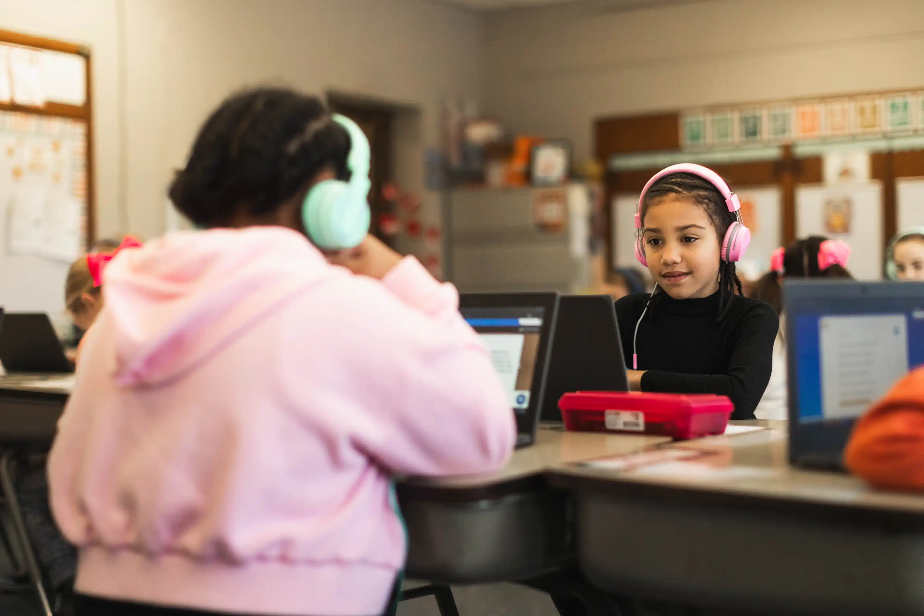 Two students wearing headphones and using laptops while seated at desks in a classroom.