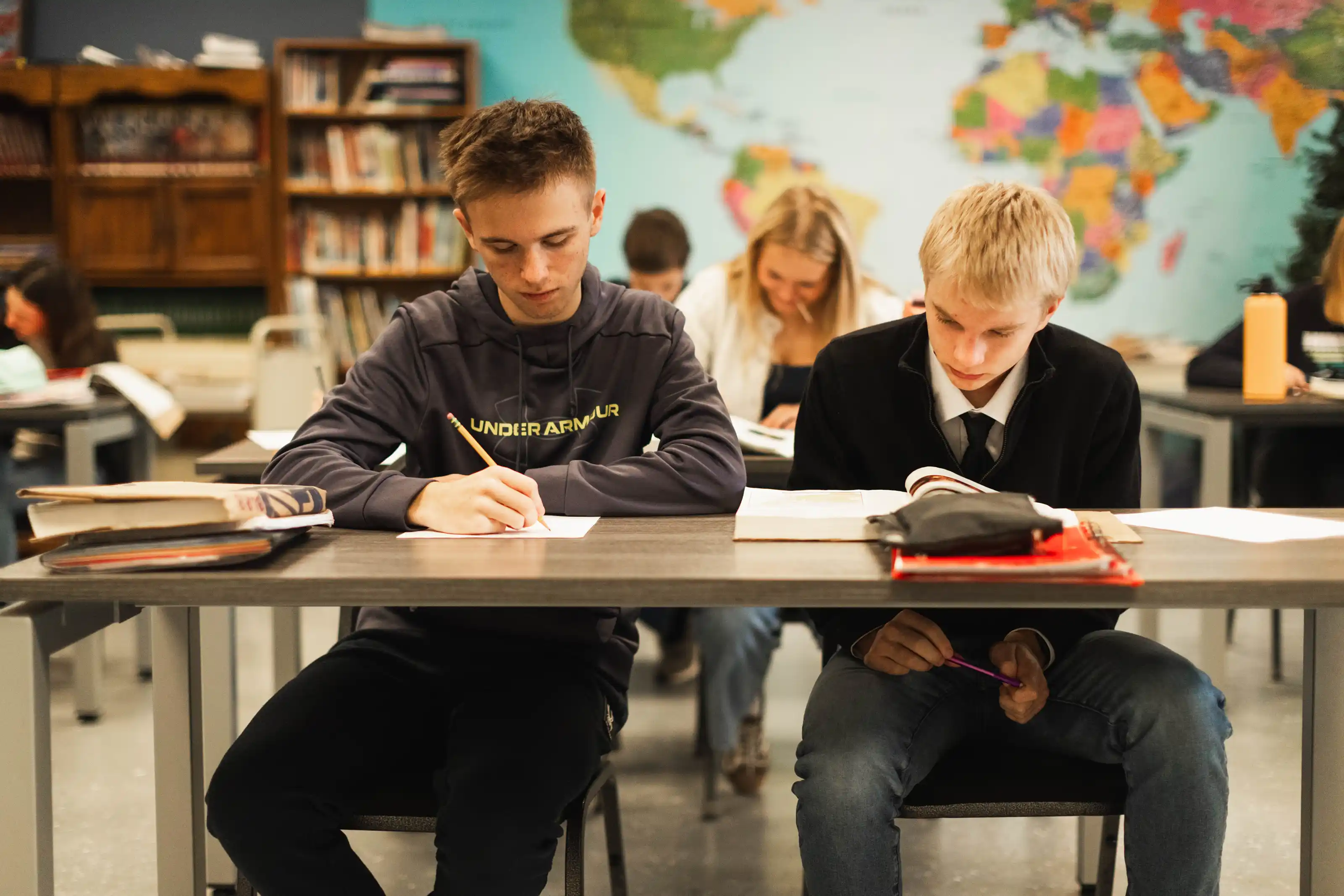 Two teenage boys sitting at a classroom desk studying; one writing with a pencil, the other reading a book with a world map on the wall behind them.