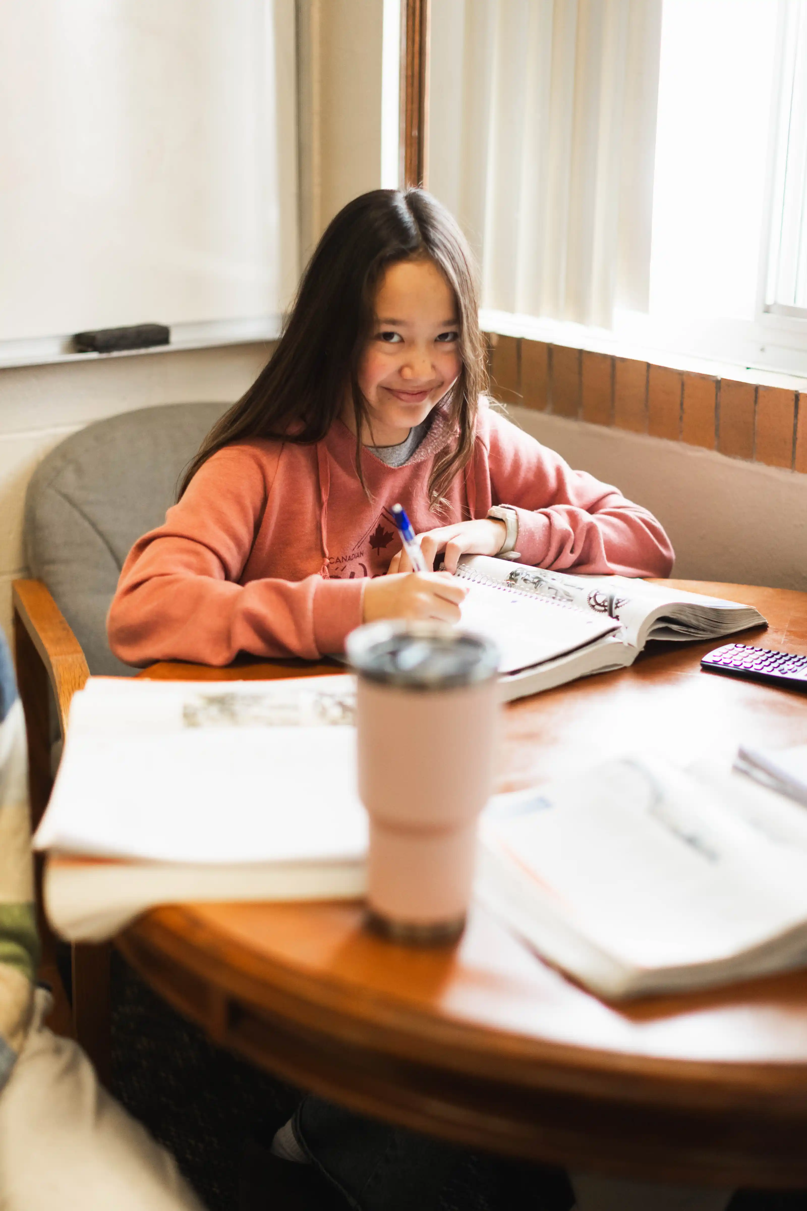 Smiling girl in a pink hoodie writing in a notebook at a round wooden table with books and a tumbler.