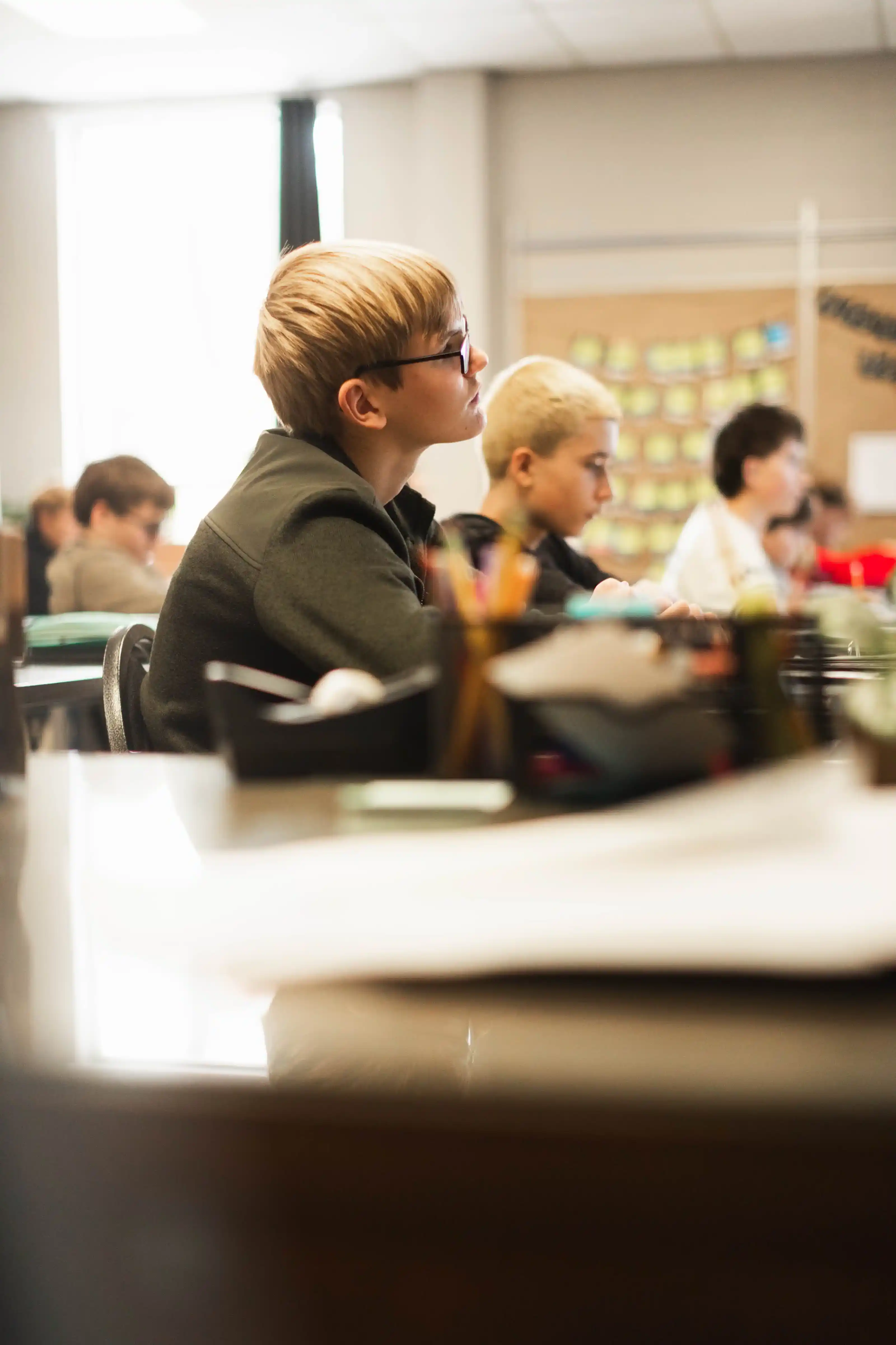 Students sitting attentively in a classroom, focusing on a lesson with blurred classroom supplies in the foreground.