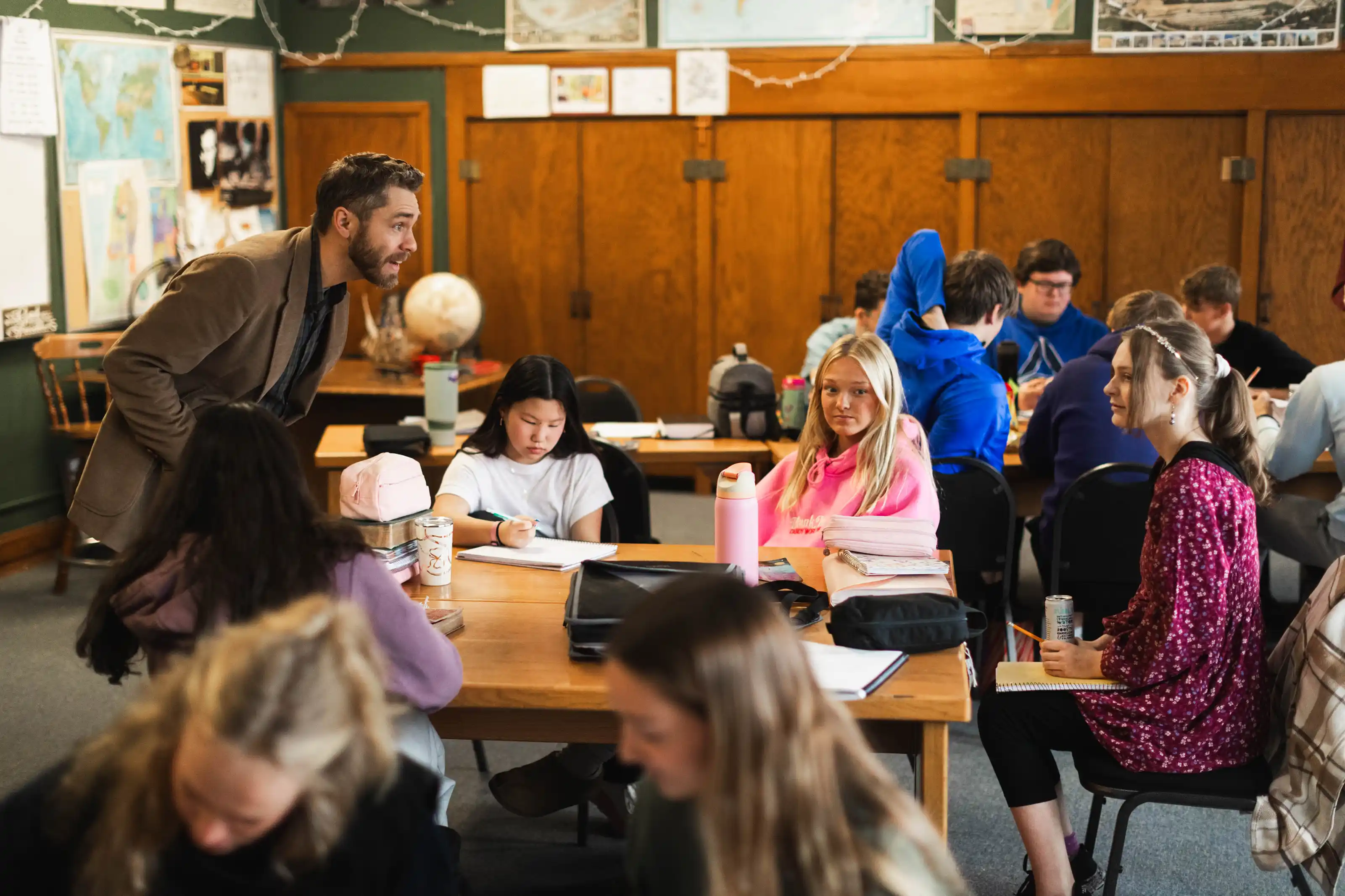 Teacher leaning over a group of students seated at tables in a classroom with wooden paneling and maps on the walls.