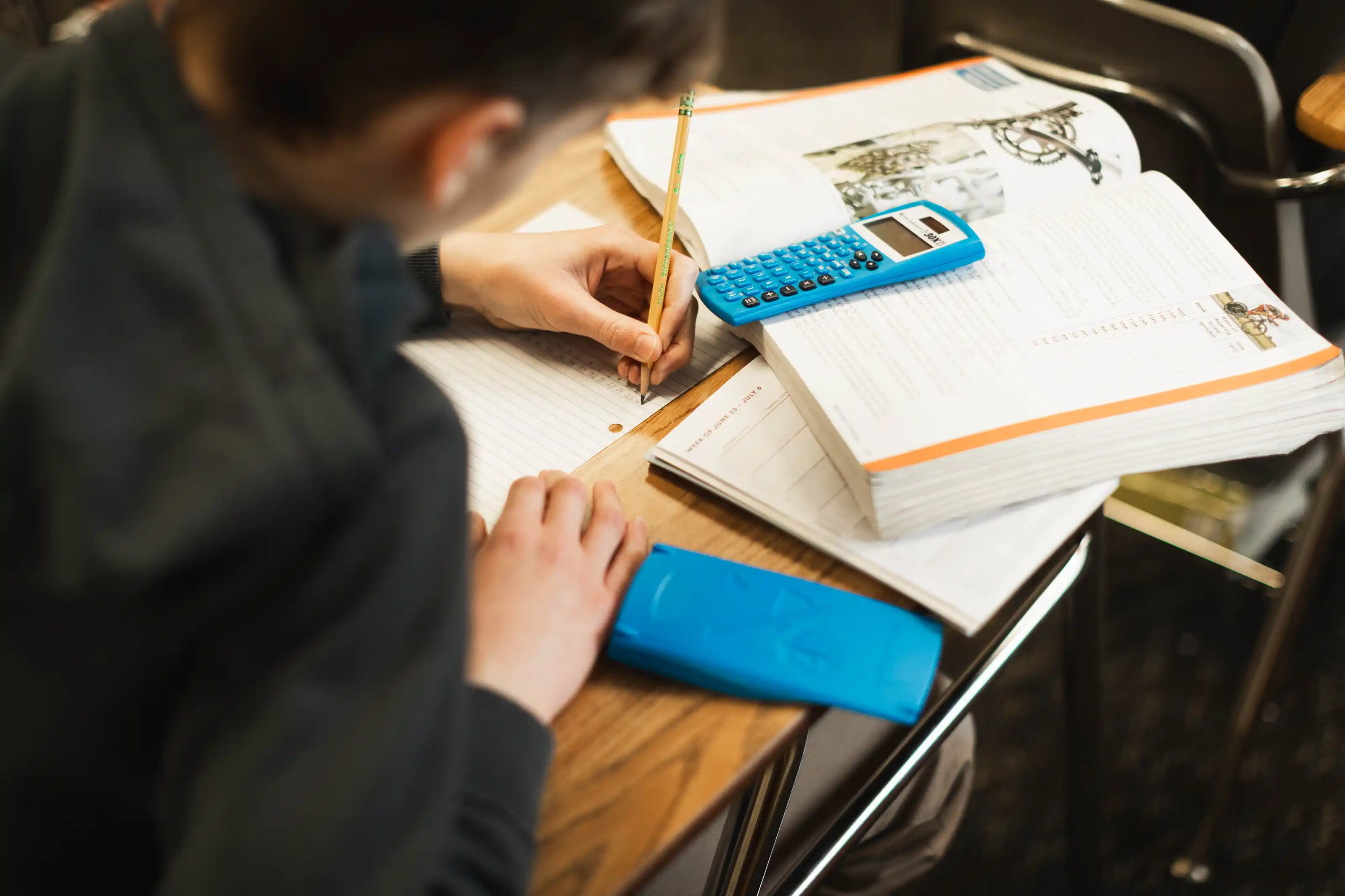 Student writing on lined paper at a desk with an open textbook and two blue calculators nearby.