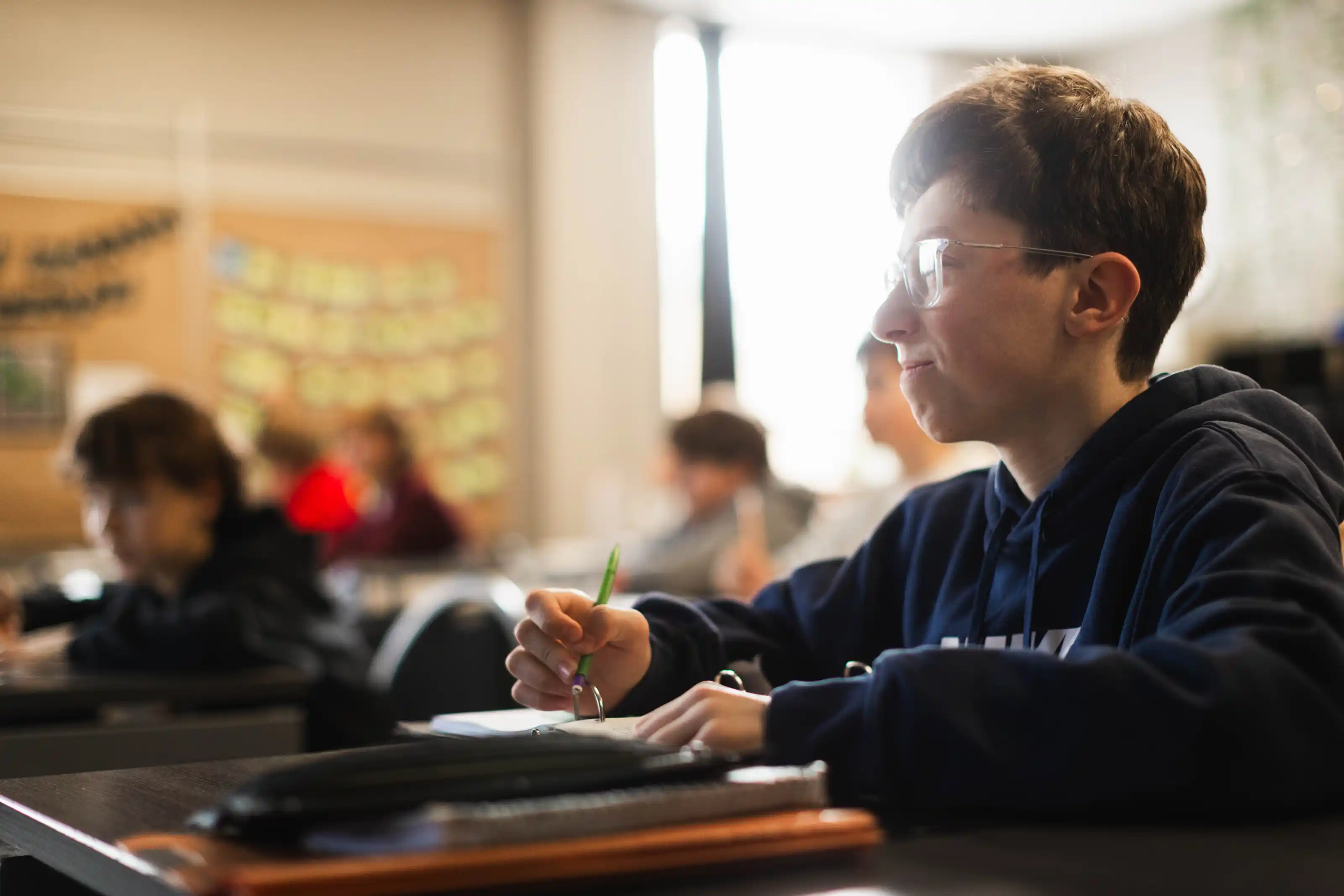 Student wearing glasses and a dark hoodie writing with a green pencil in a classroom.