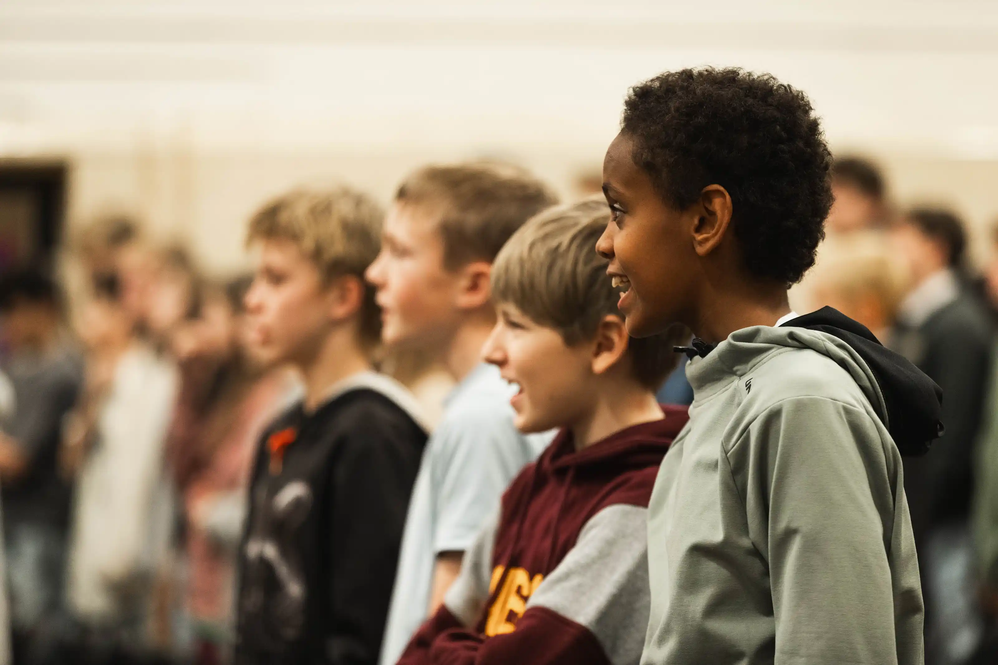 Four boys standing in a line indoors looking attentively to the right, with the boy in the front smiling.