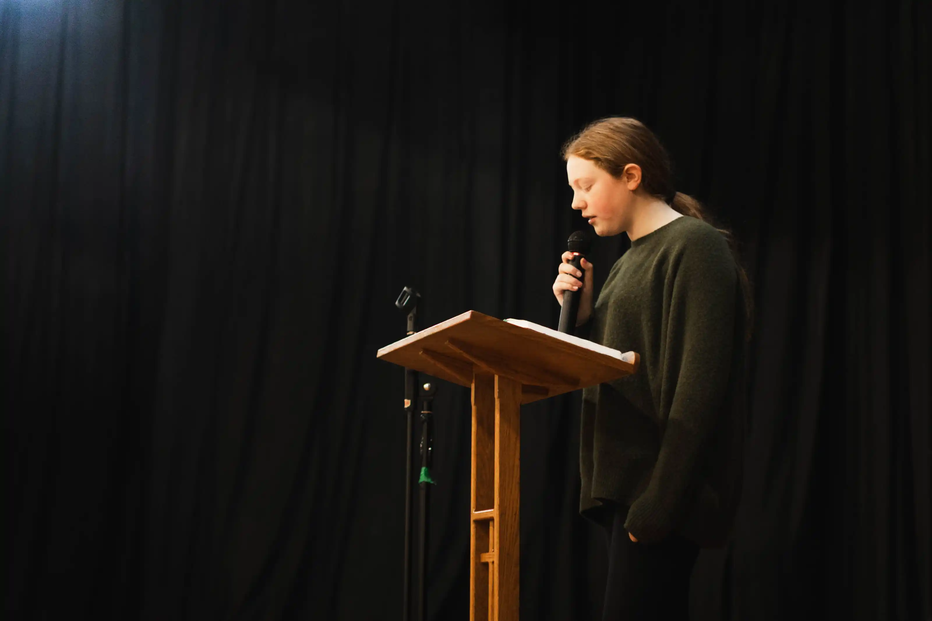 Young woman in a dark sweater speaking into a microphone at a wooden podium against a black curtain background.