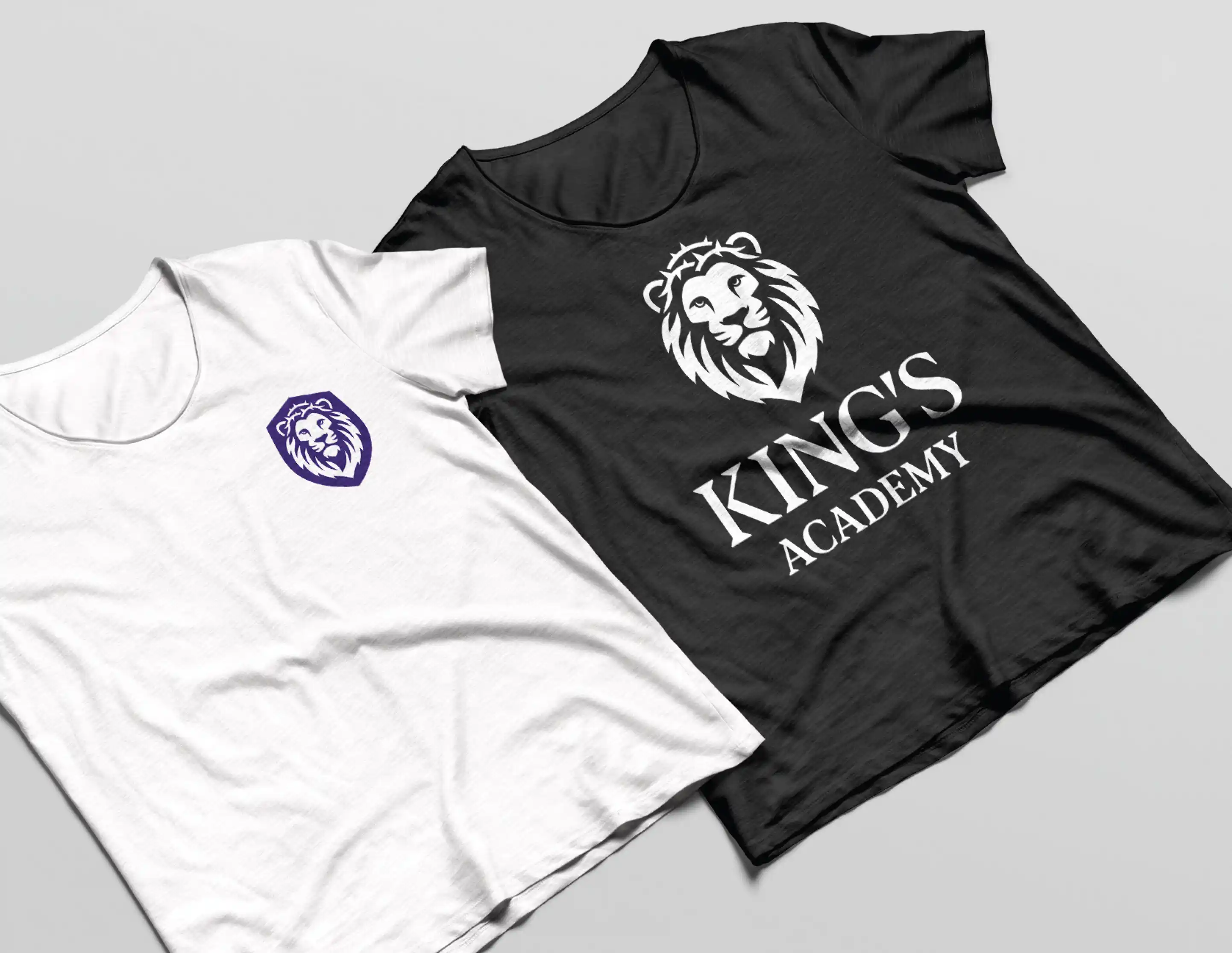 Two t-shirts, one white with a purple lion head logo on the chest and one black with a large white lion head and the text 'KING'S ACADEMY'.