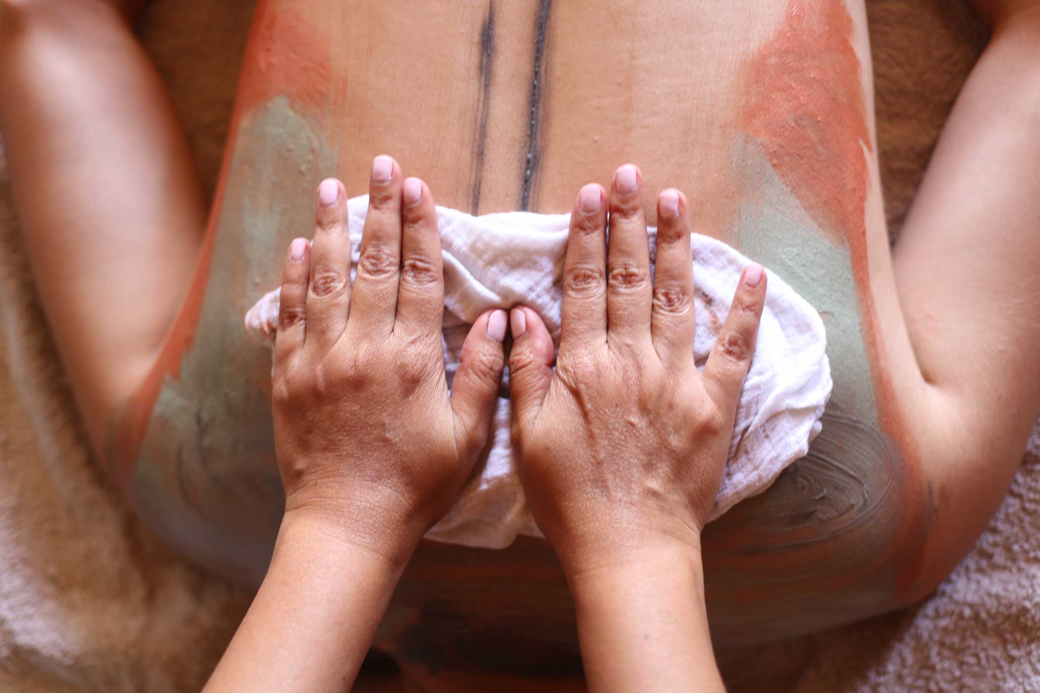 Mains appliquant une serviette blanche sur le dos enduit d'argile de différentes couleurs d'une personne allongée pour un soin spa.
