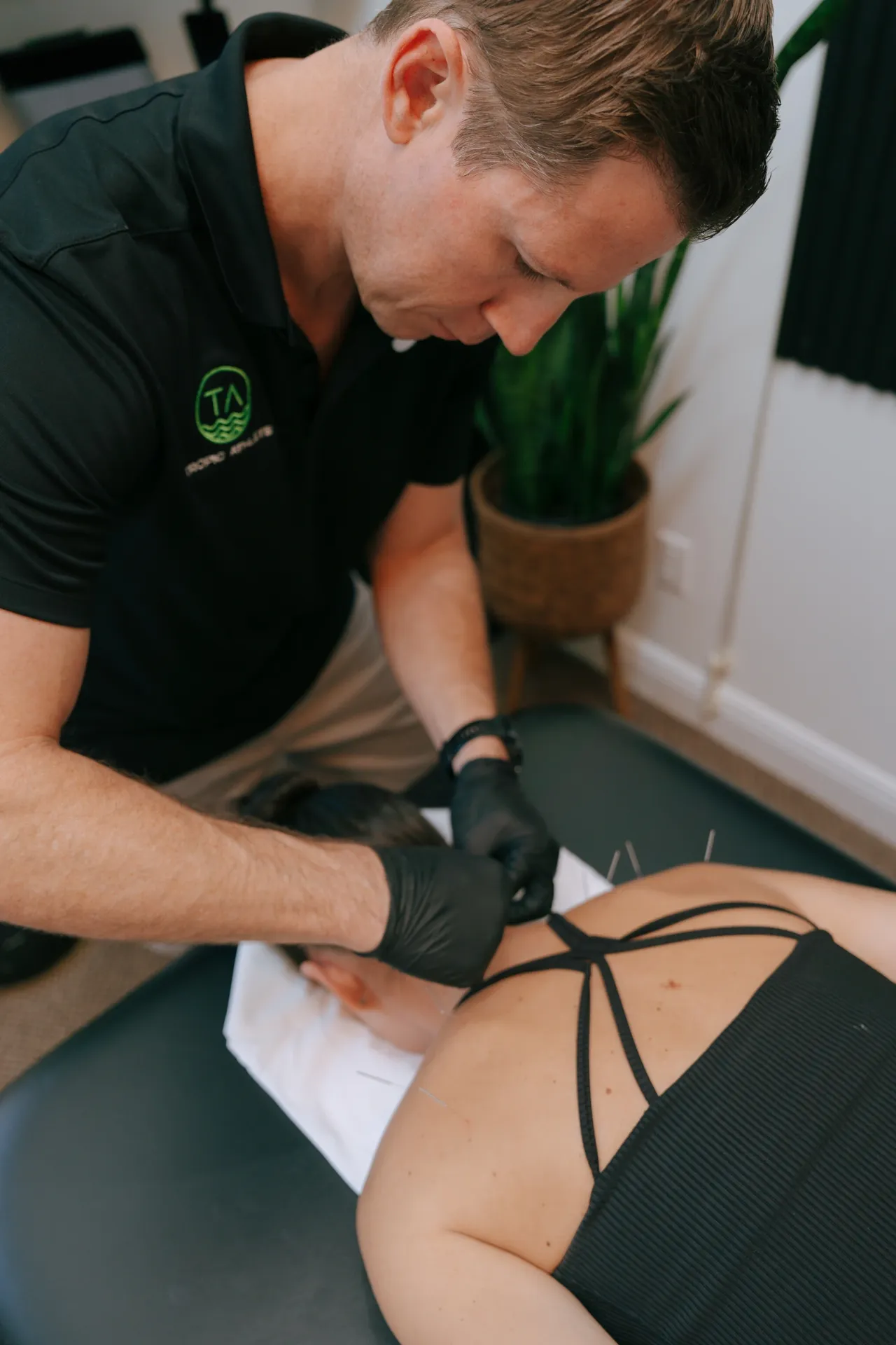 Therapist wearing black gloves performing acupuncture on a woman's upper back in a treatment room. Tropic Athlete