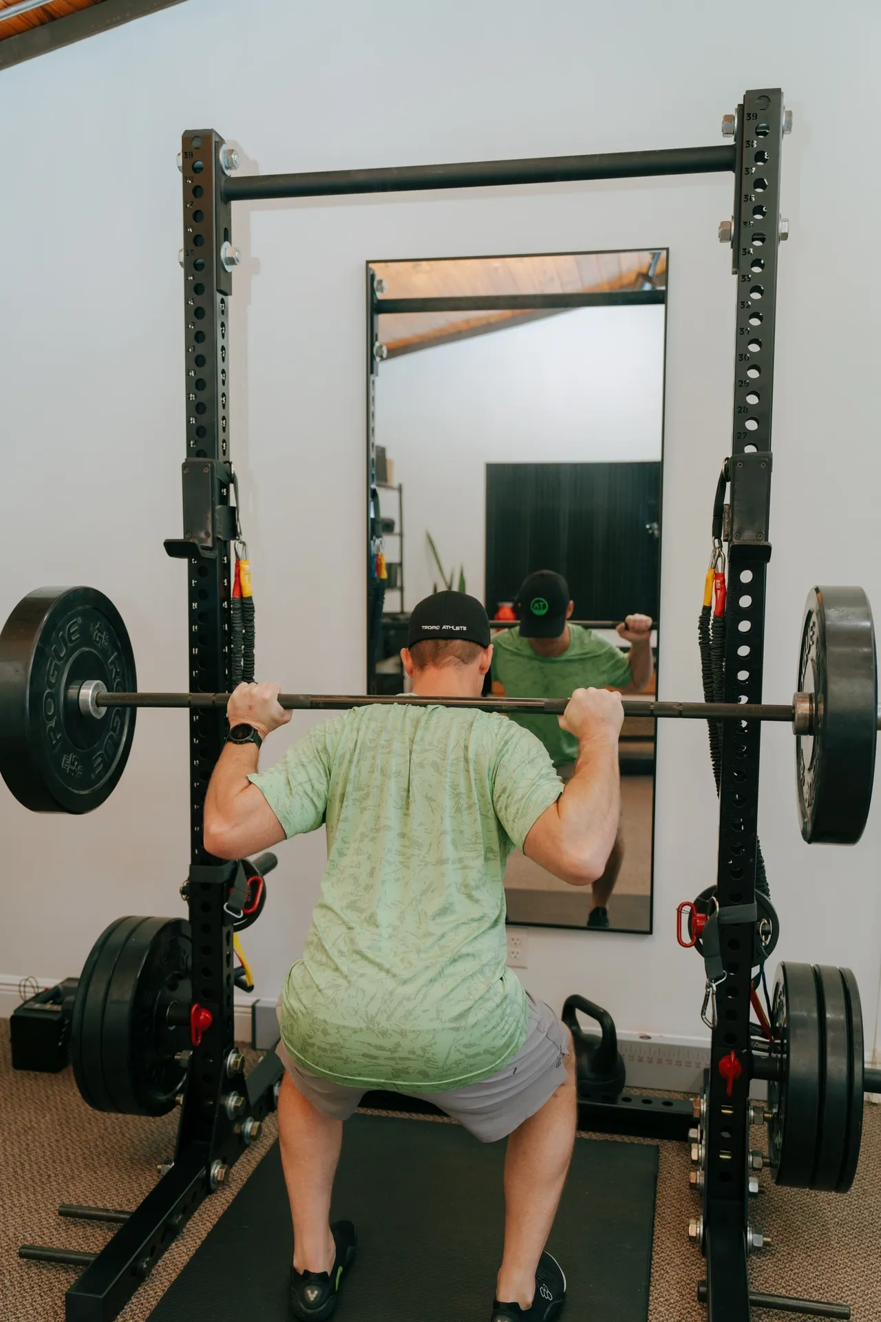 Man in a green shirt and black cap doing a barbell squat in front of a mirror in a home gym. Tropic Athlete