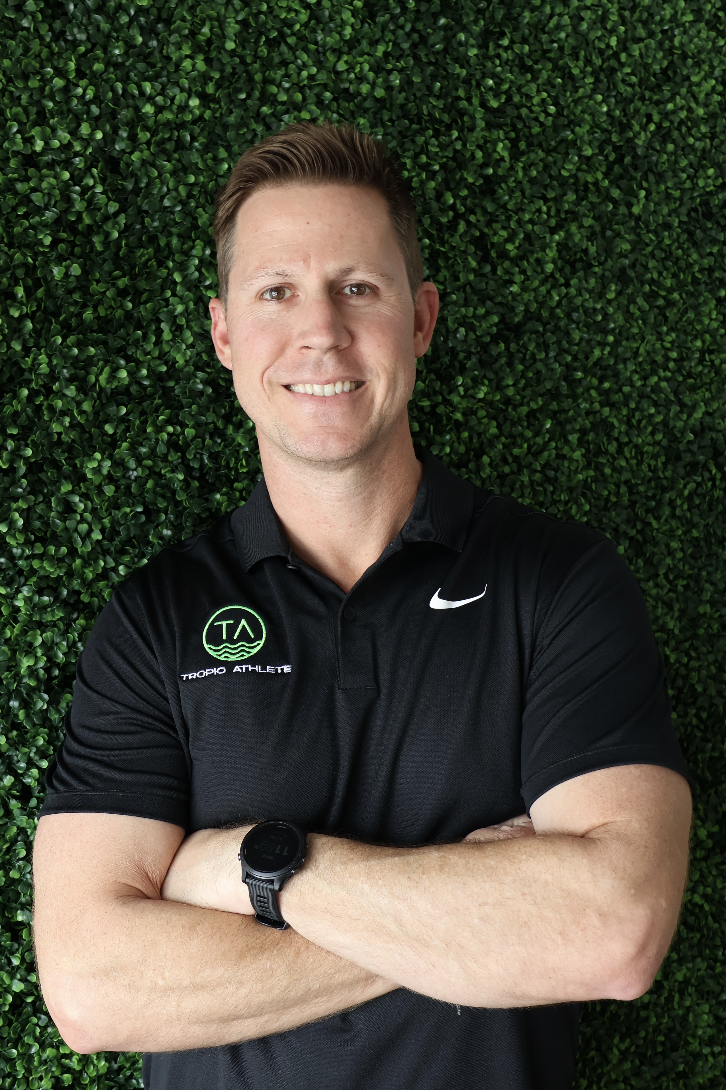 Smiling man with short brown hair wearing a black polo shirt with Tropio Athlete logo, standing with arms crossed against a green leafy background. Tropic Athlete