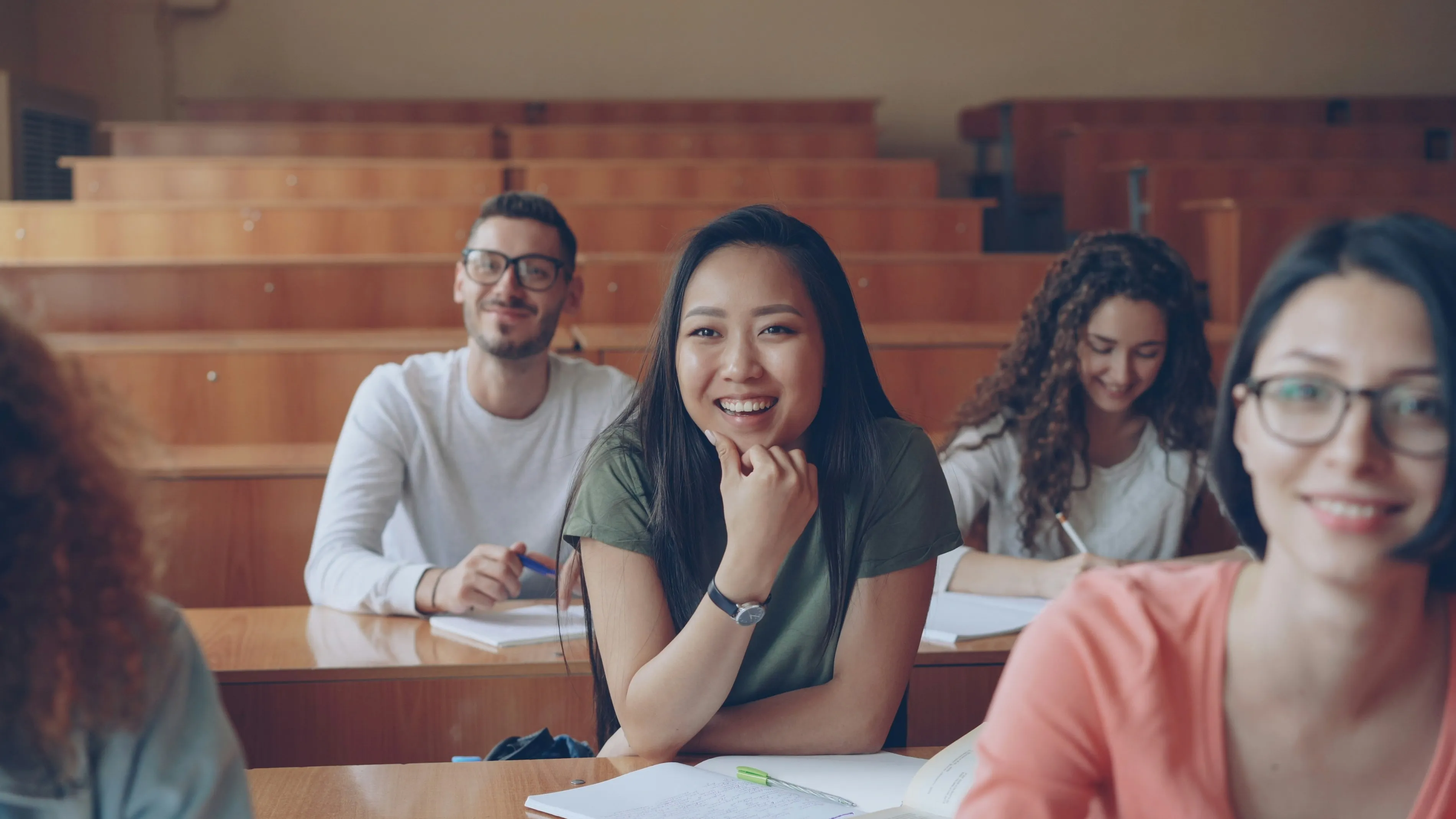 A diverse group of four college students smiling and seated in a classroom with wooden desks.