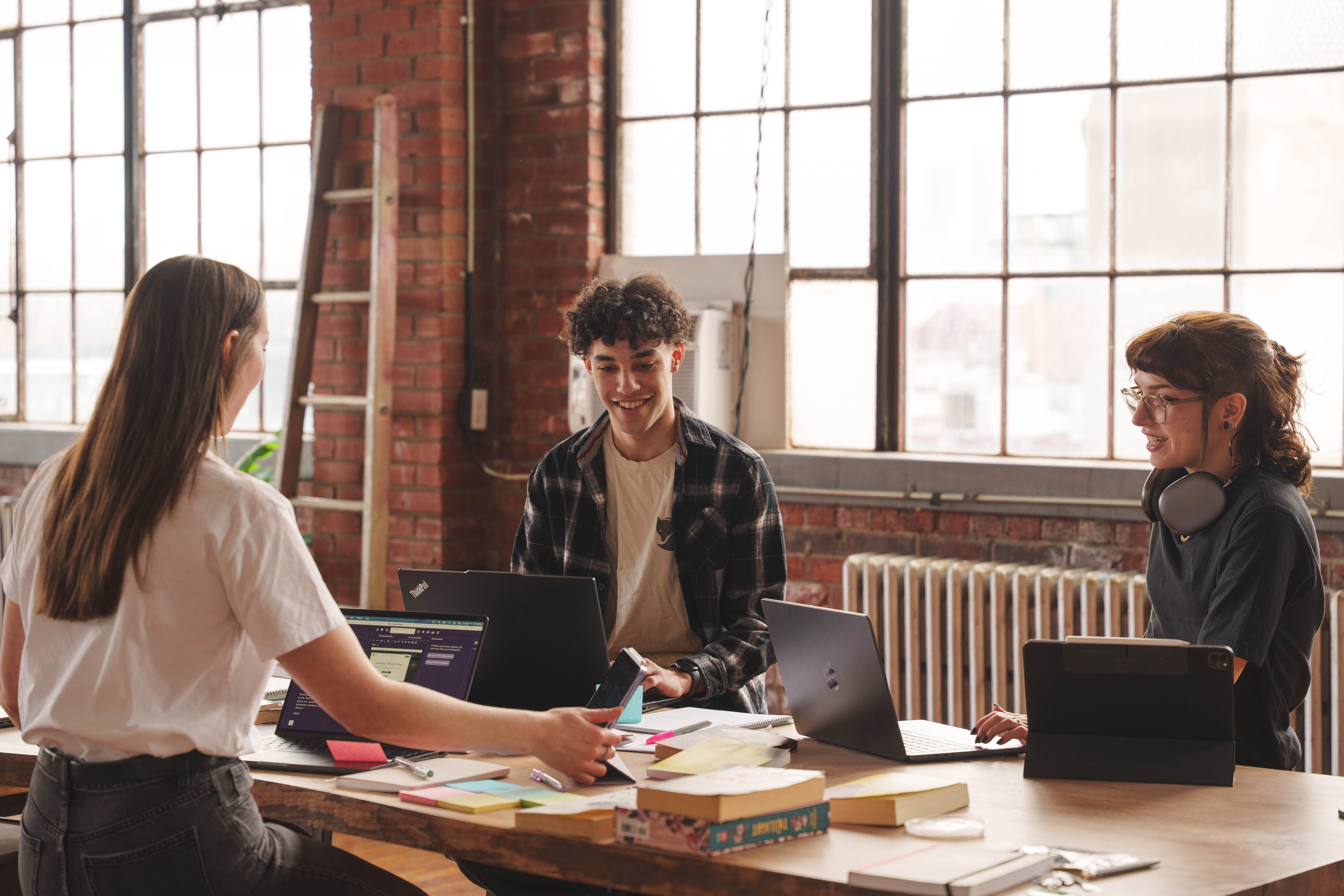 Three young adults studying with laptops and books around a wooden table in a bright room with large windows and exposed brick walls.