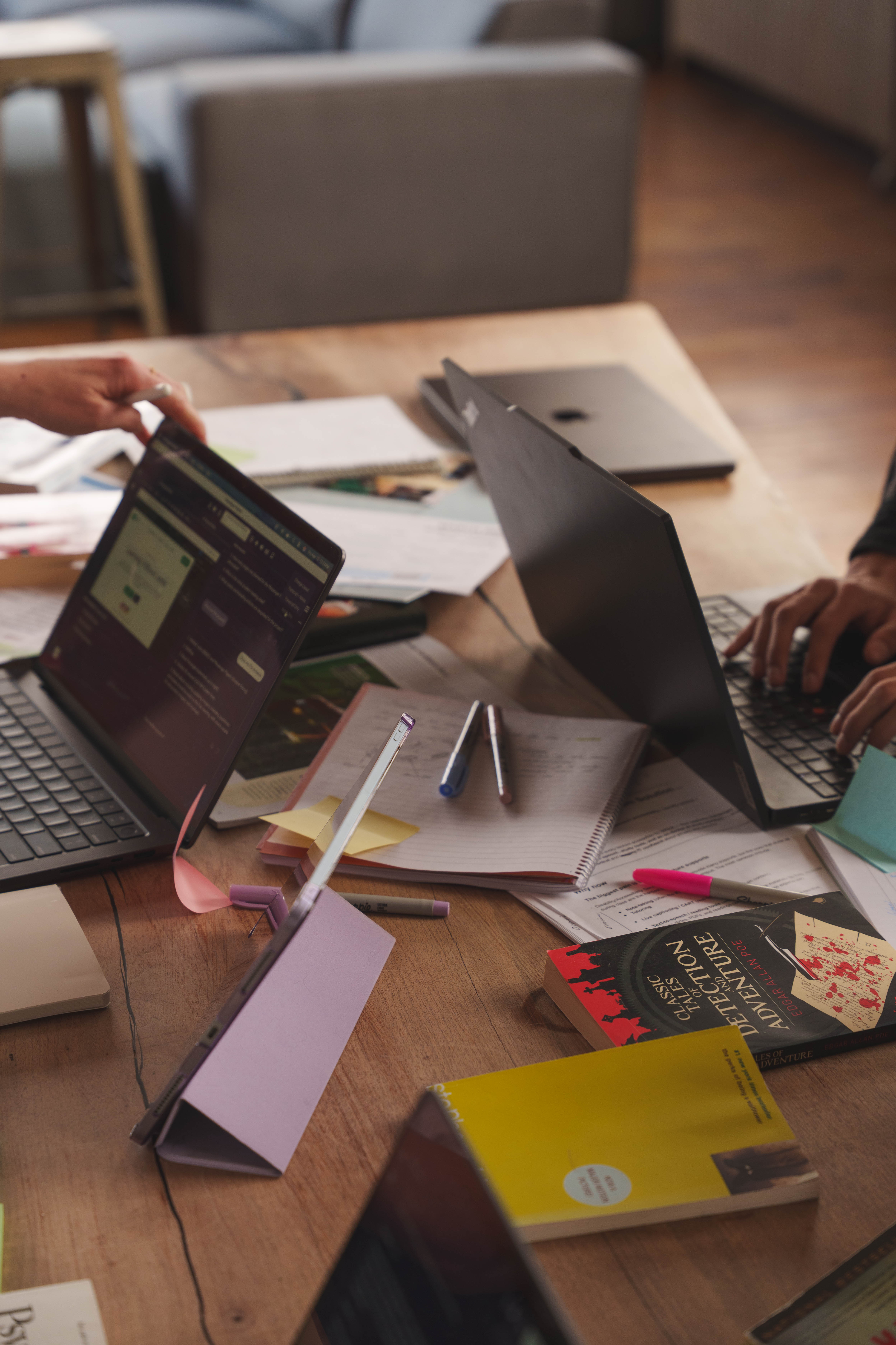 Wooden table with open laptops, notebooks, pens, papers, and books including 'Classic Tales of Detection and Adventure' during a study session.