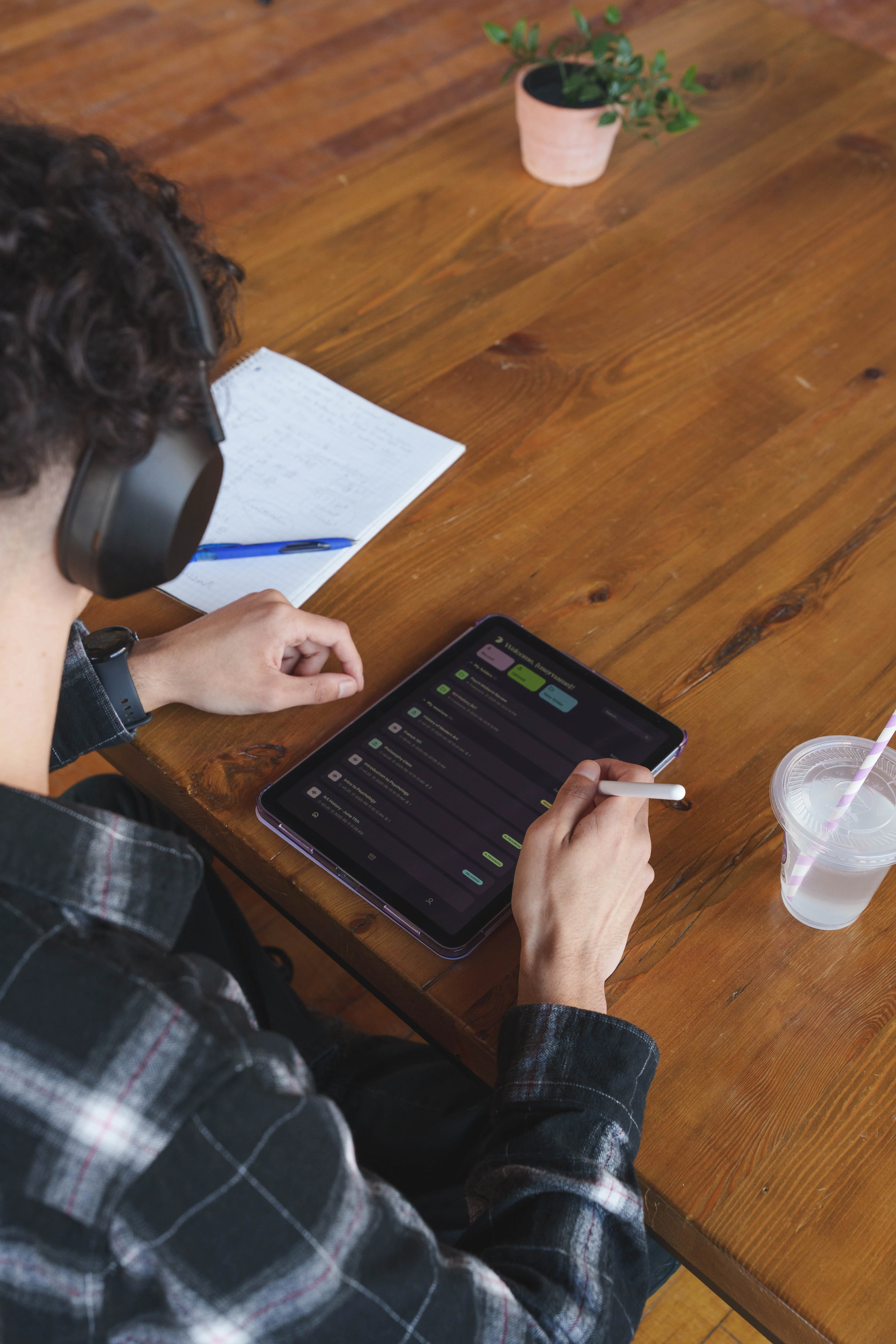 Person wearing headphones using a stylus on a tablet displaying the Messenger Pigeon app while sitting at a wooden table with a notebook, pen, plant, and plastic cup with a straw.