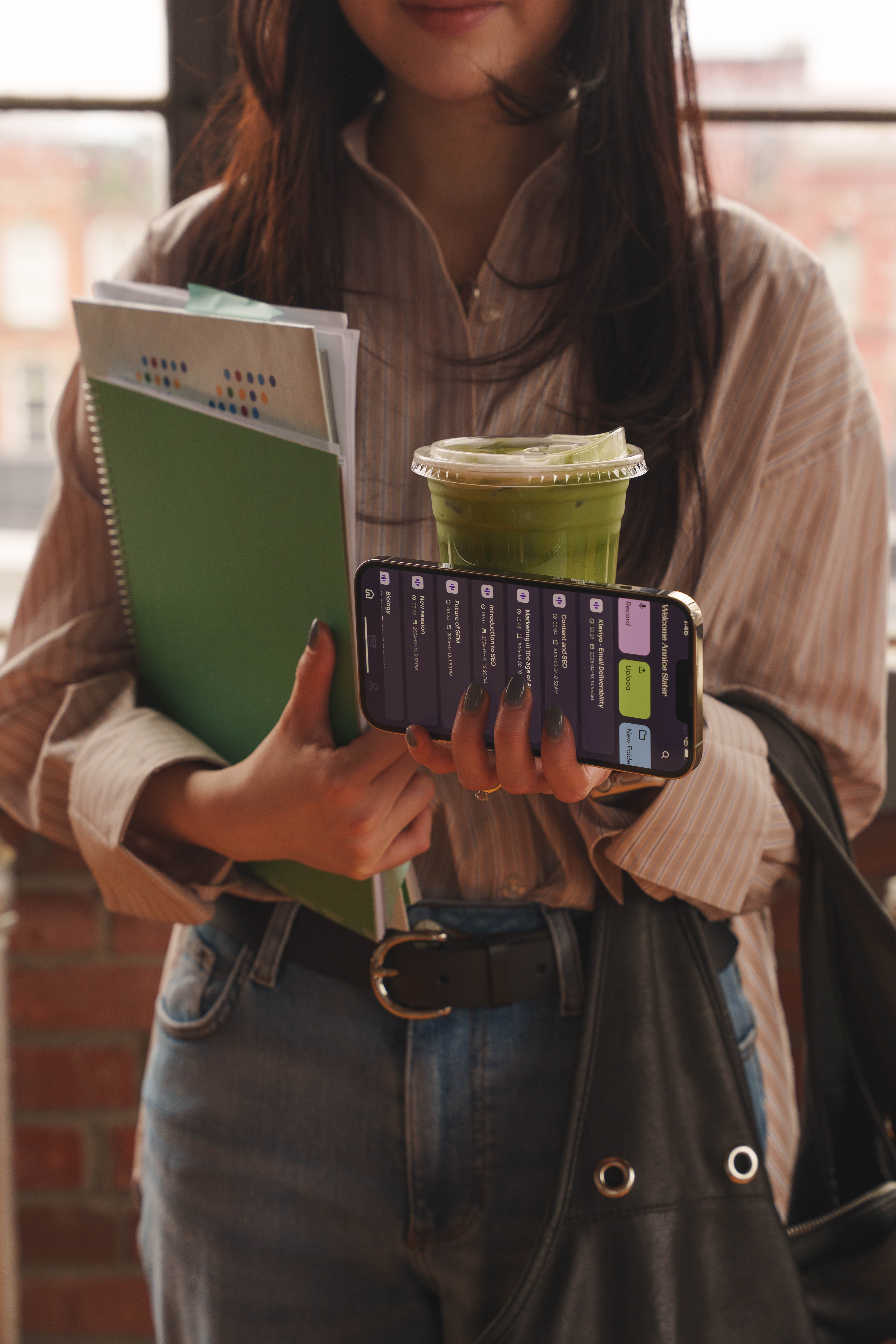 Person holding notebooks, a smartphone displaying Messenger Pigeon App, and a green iced drink with a plastic lid.
