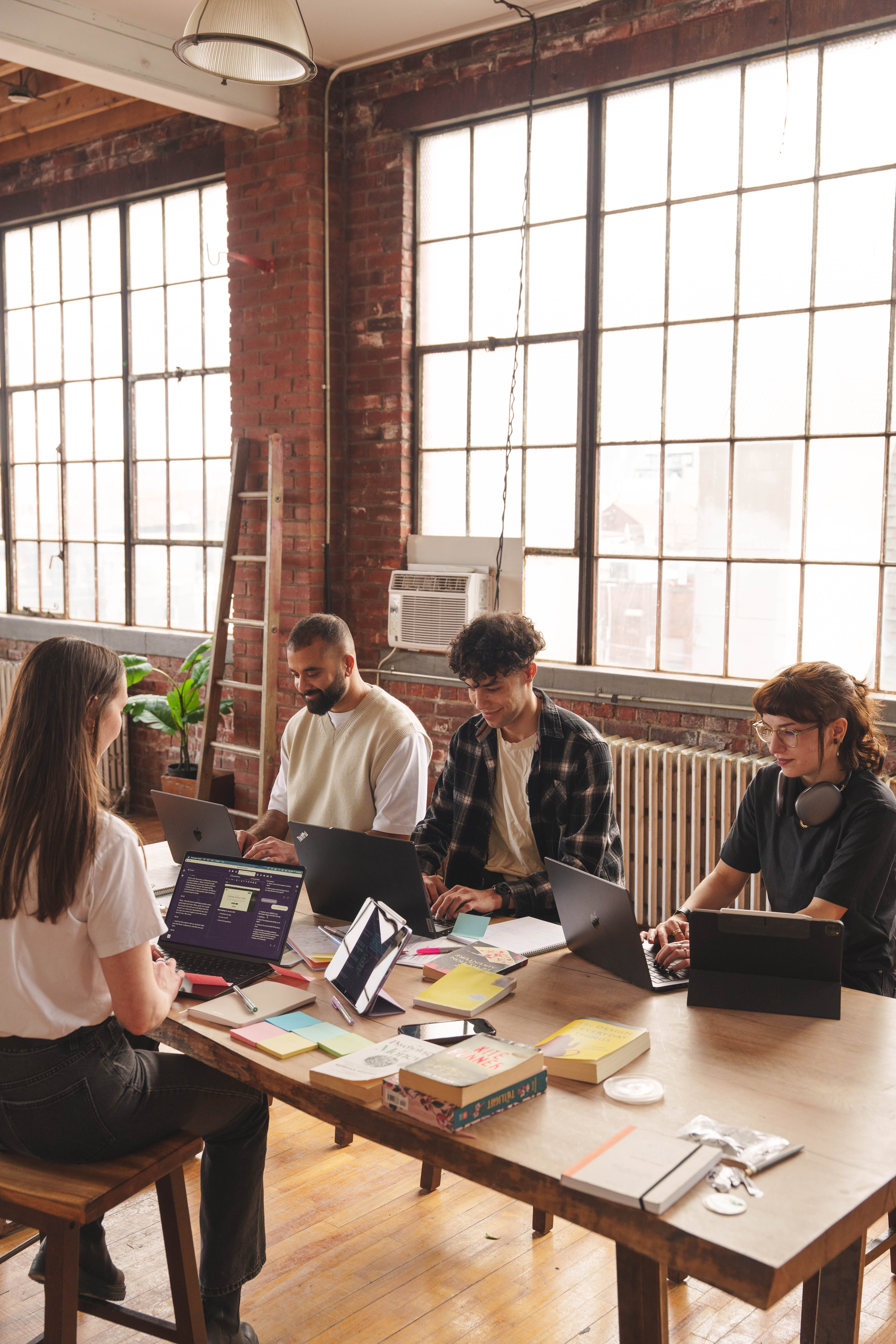 Four diverse people sitting around a wooden table in a bright industrial-style room, working on laptops with books, notebooks, and sticky notes scattered on the table.