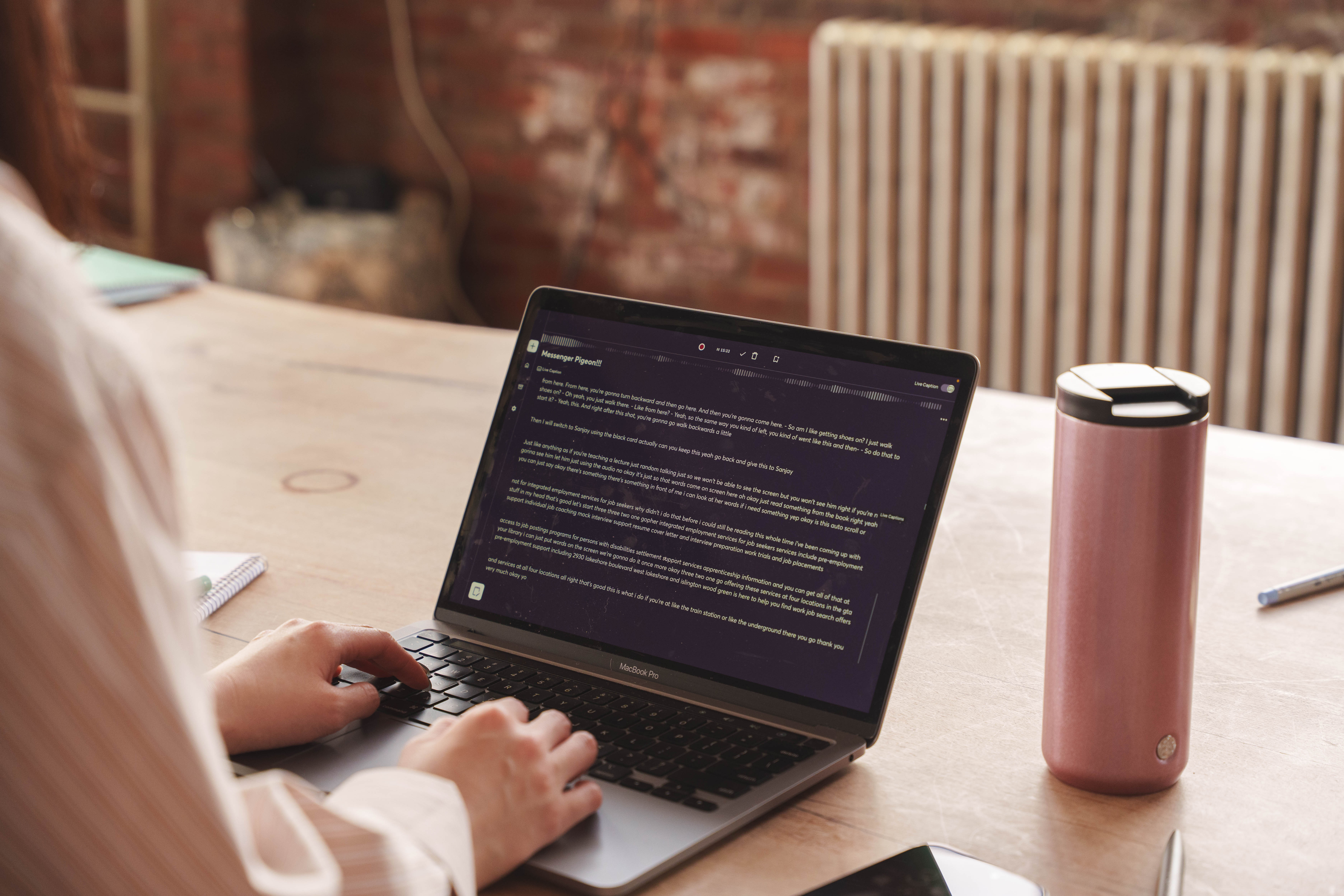 Person typing on a MacBook Pro laptop displaying a live captions and transcripts on a wooden table with a pink insulated bottle nearby.