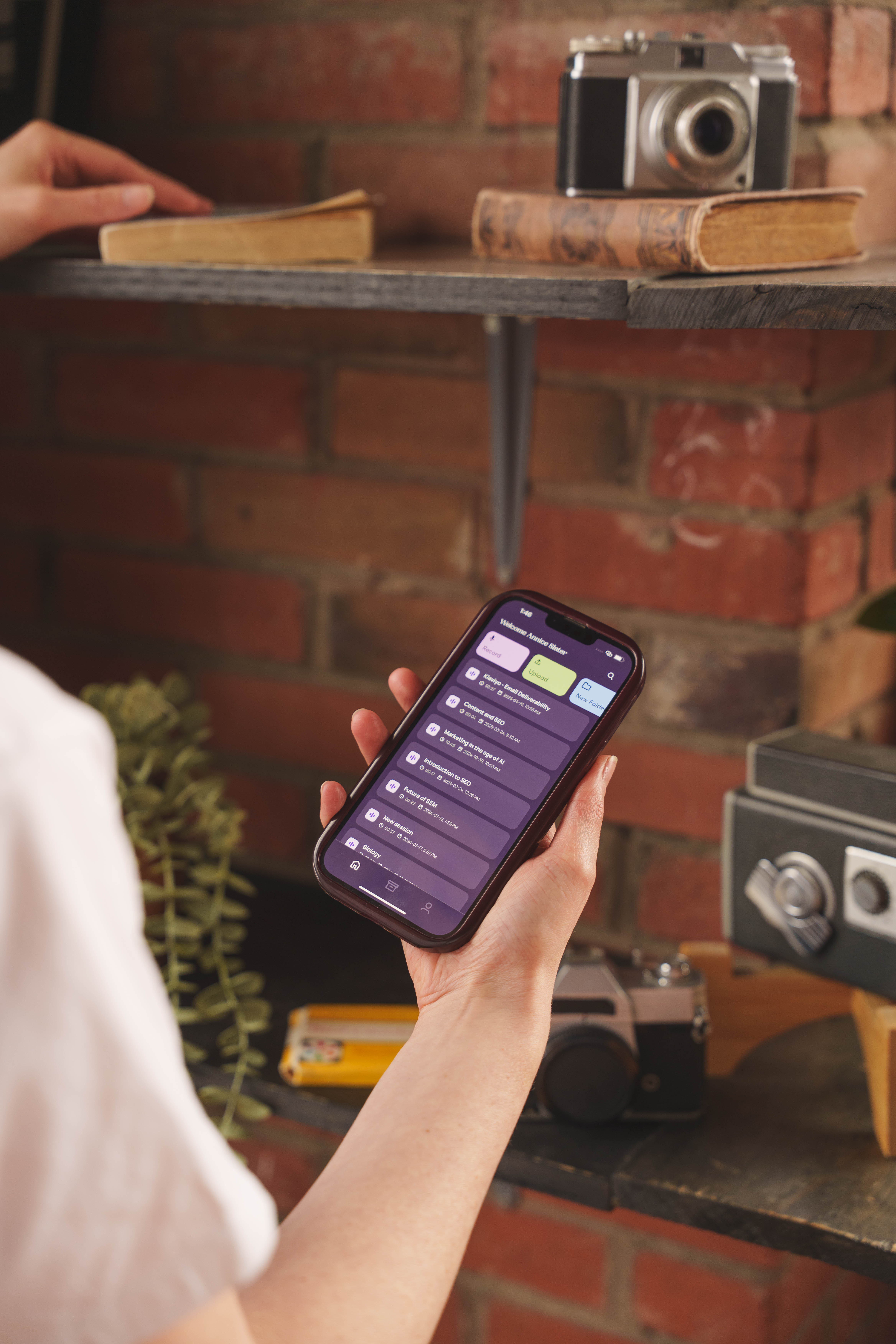 Person holding a smartphone showing a recording app interface with a list of audio files against a brick wall background with shelves holding vintage cameras and books.