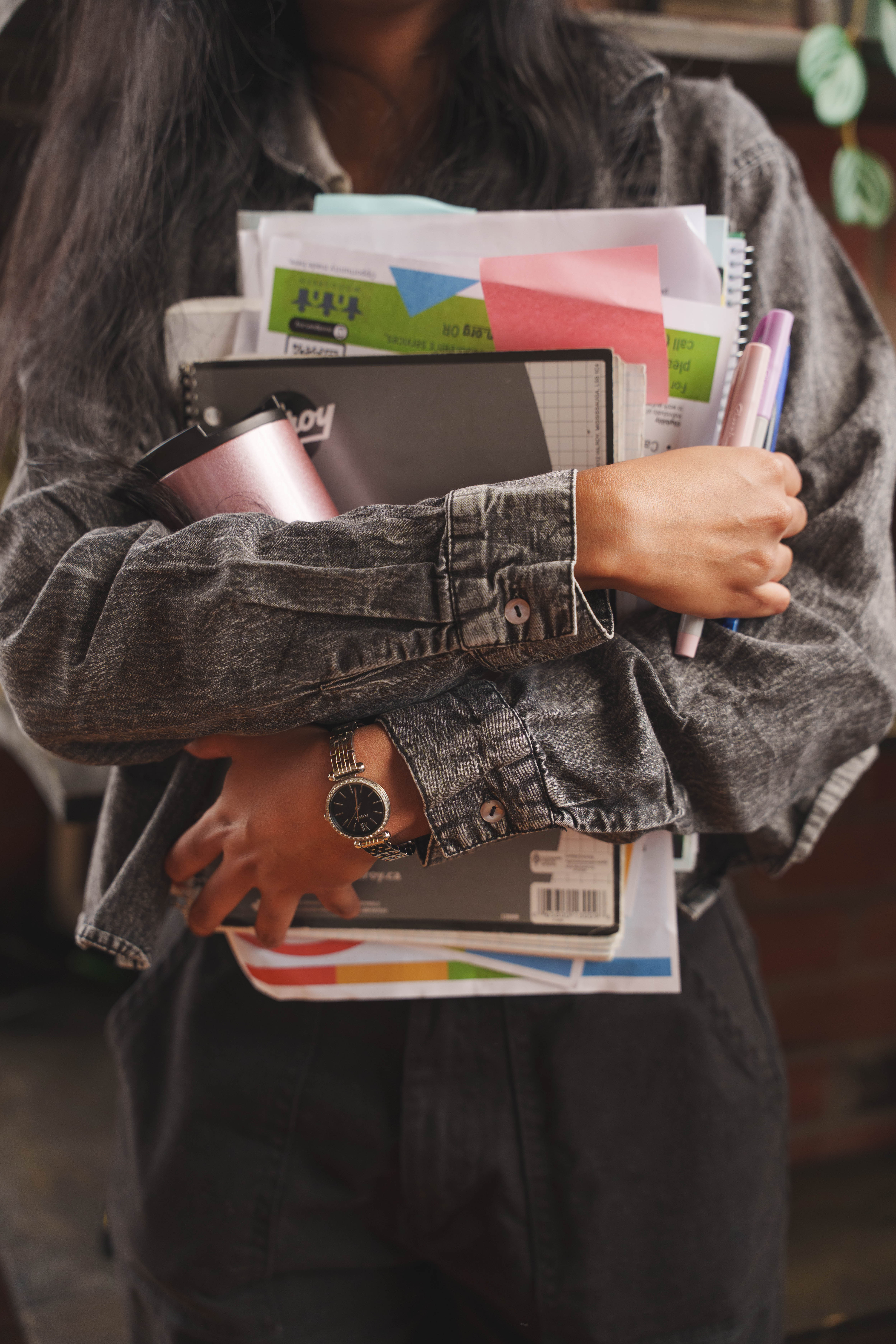 Person wearing a grey denim shirt holding notebooks, papers, pens, and a pink tumbler against their chest.