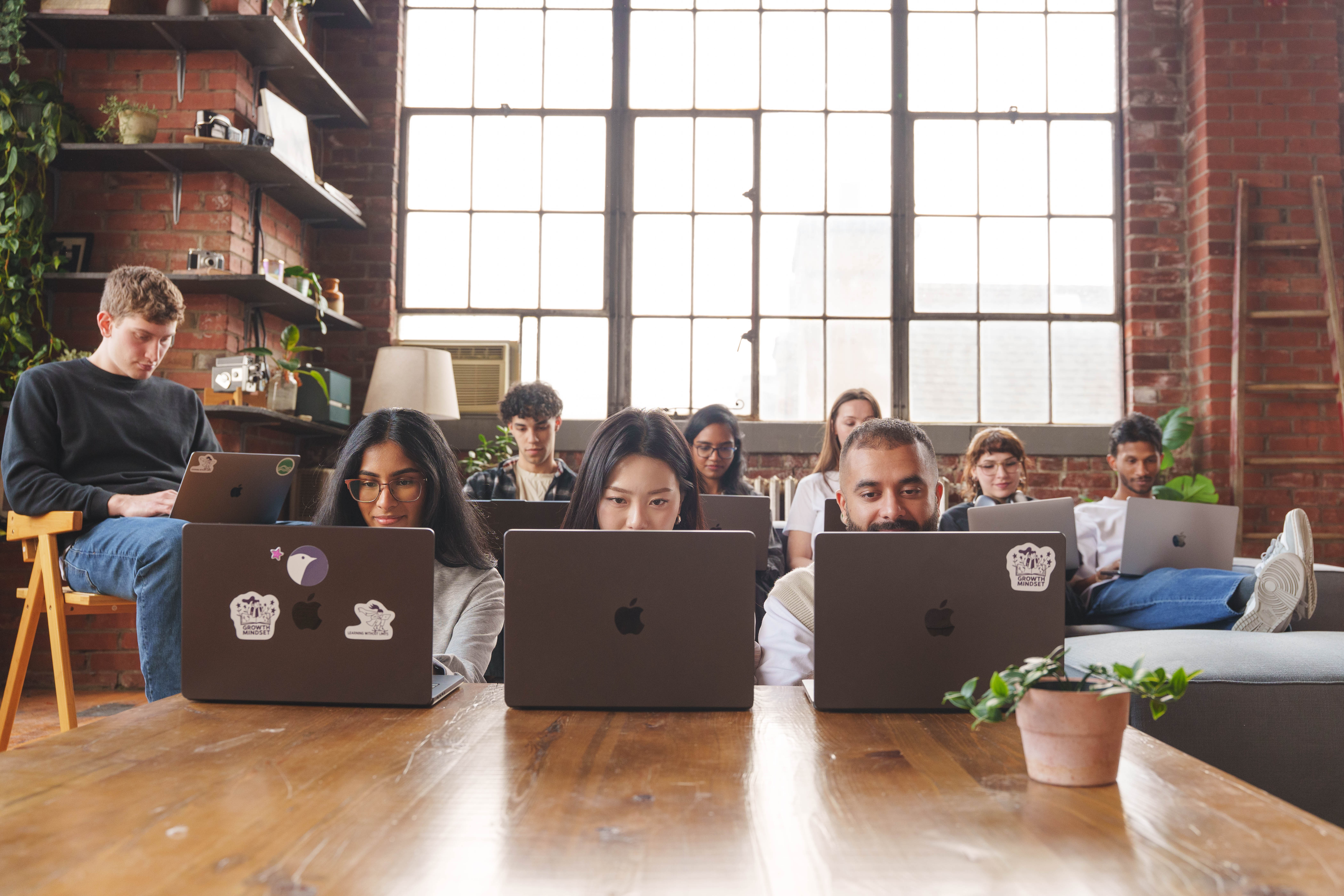 Group of diverse young adults working on laptops in a bright room with large windows and brick walls.