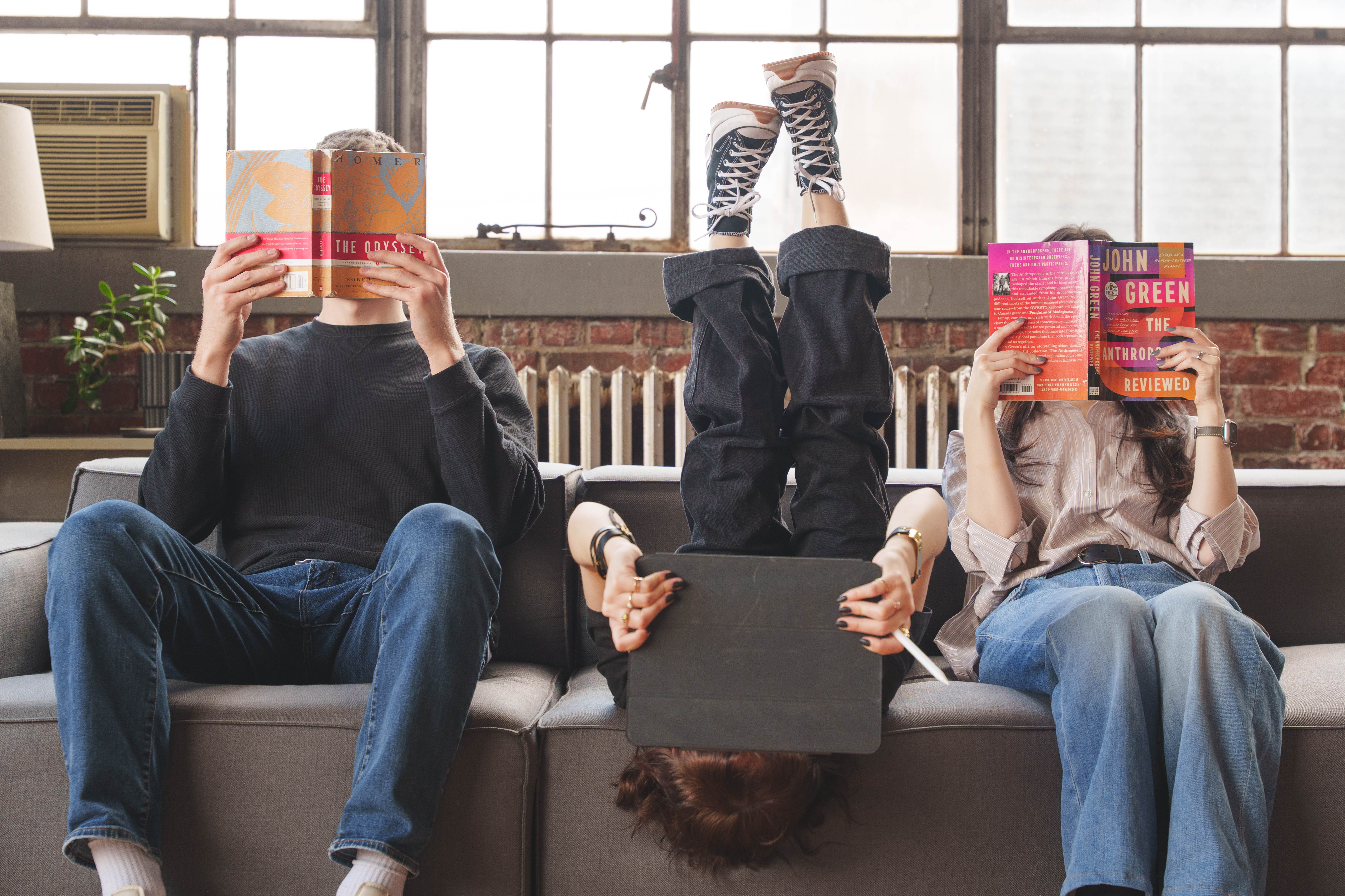 Three people on a couch engaged in reading, two holding books and one holding a tablet upside down with legs raised.