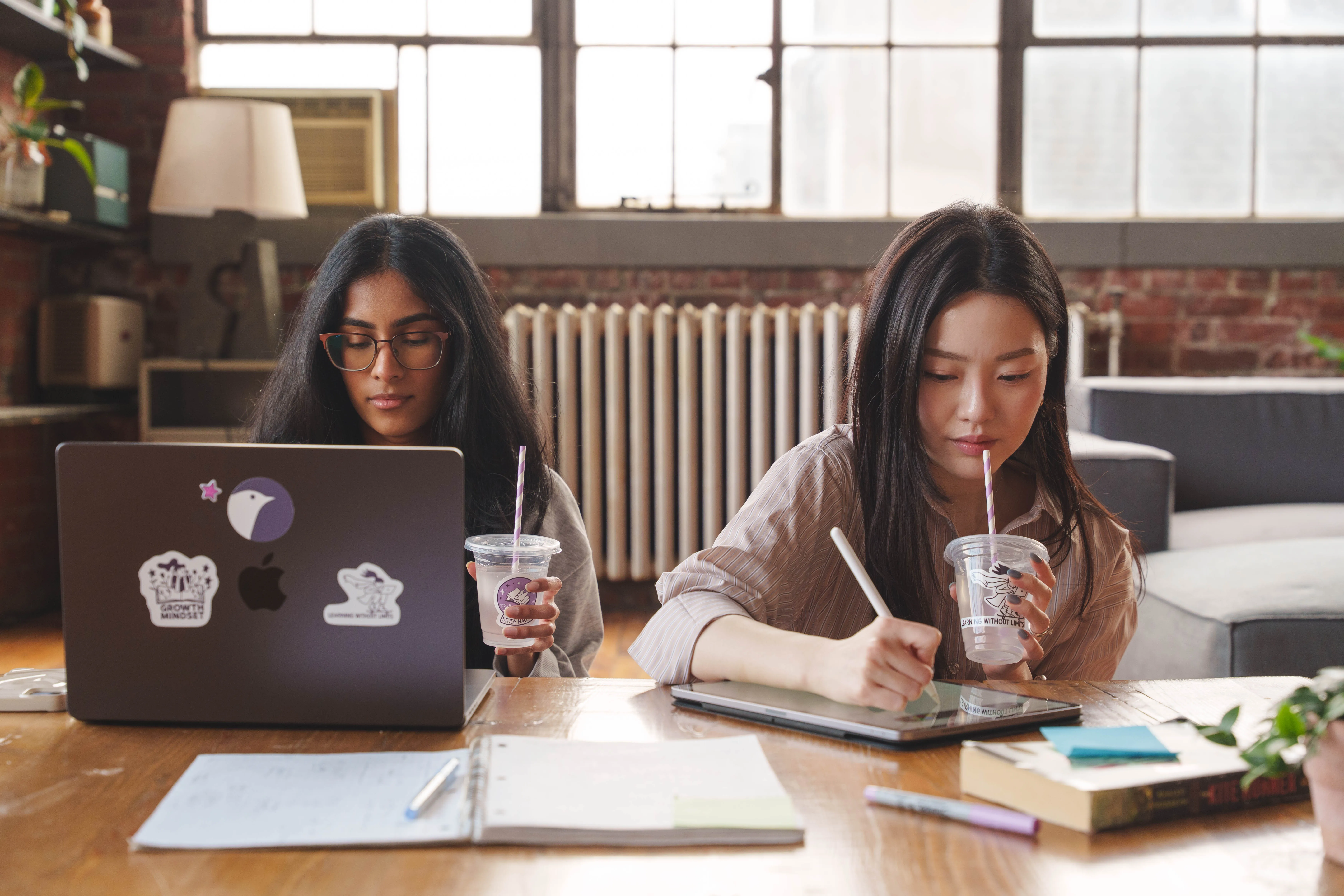 Two women studying at a wooden table, one using a laptop and the other writing on a tablet, both holding drinks with straws.