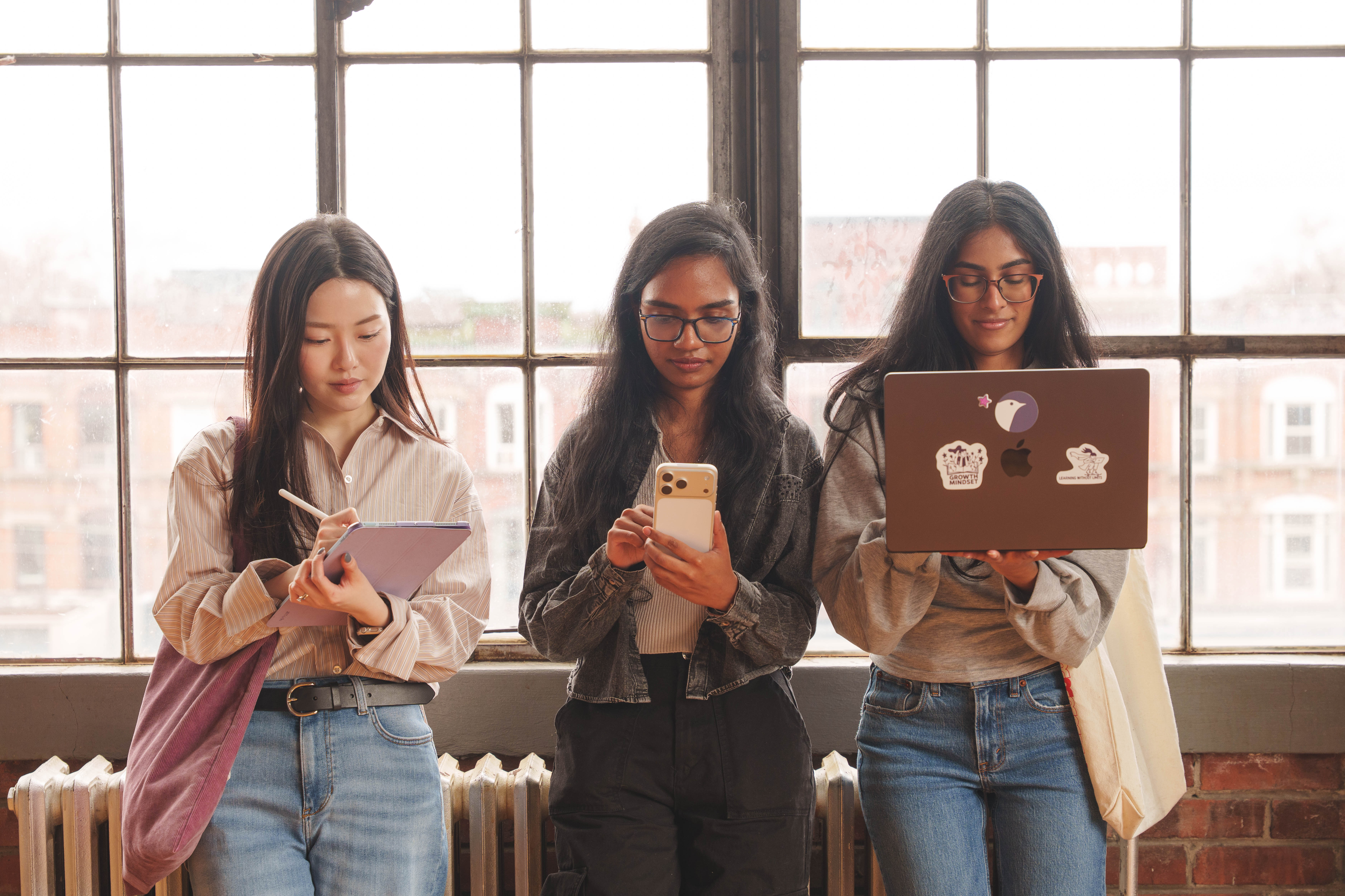 Three young women standing in front of a large window using digital devices: one writing on a tablet, one holding a smartphone, and one working on a laptop with stickers.