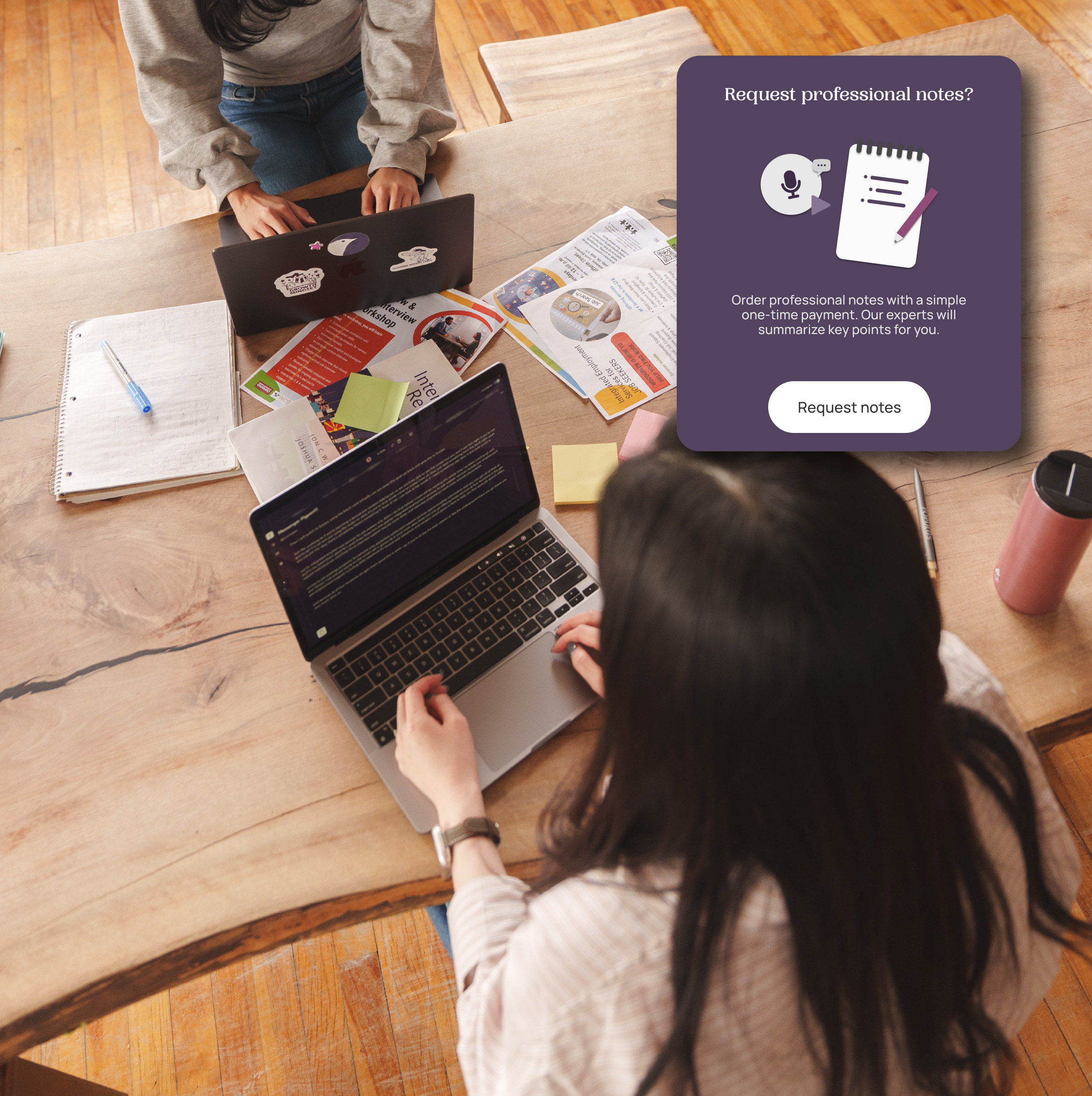 Two people working on laptops at a wooden table with papers, notebooks, and sticky notes scattered around.