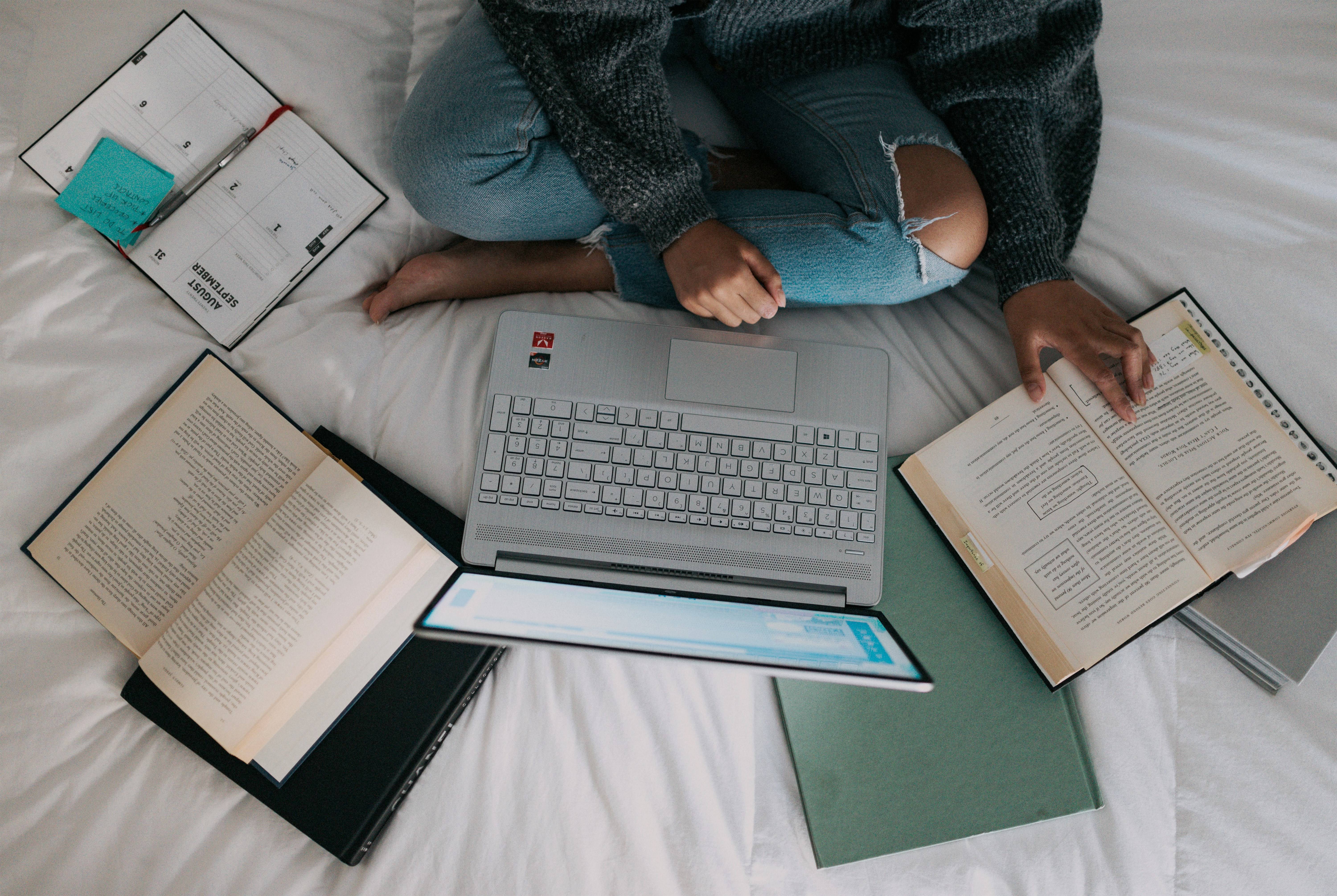 A person, crossed legged on a bed, with a laptop and books surrounding them.