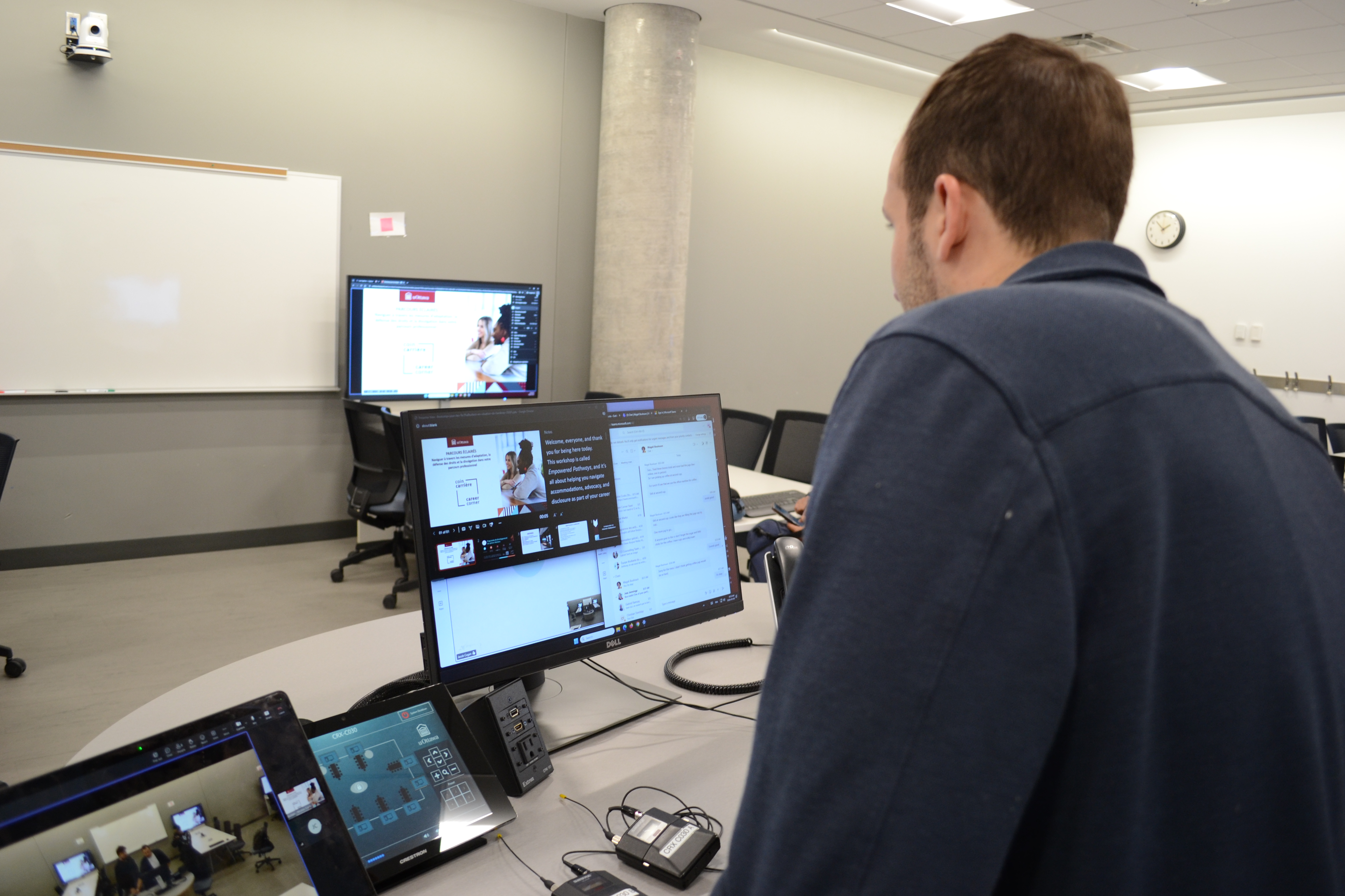 Person operating classroom presentation and live captions technology while monitoring a live hybrid session in a university classroom.