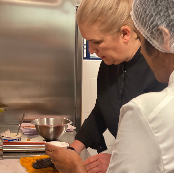 Une cheffe souriante en tenue blanche dresse une table élégante en extérieur avec des amuse-bouches, des plats colorés et des compositions florales, illustrant une expérience traiteur printanière raffinée et conviviale.