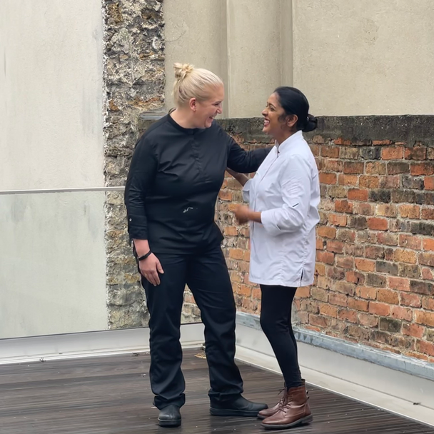 Une main verse une boisson rose rafraîchissante dans un verre sur une table élégante en extérieur, entourée de fruits, de fleurs et de plats colorés, illustrant une ambiance conviviale et estivale.