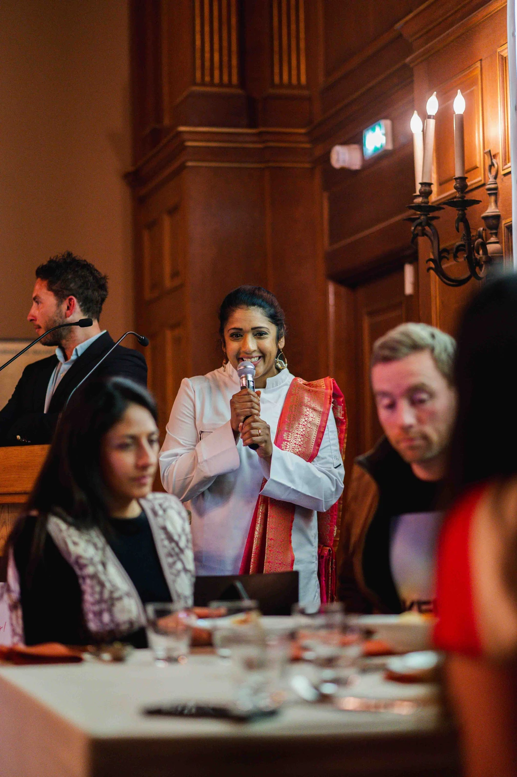 Une main verse une boisson rose rafraîchissante dans un verre sur une table élégante en extérieur, entourée de fruits, de fleurs et de plats colorés, illustrant une ambiance conviviale et estivale.