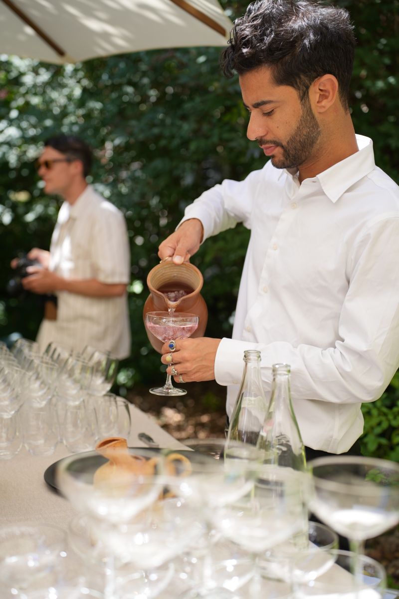 Une cheffe souriante en tenue blanche dresse une table élégante en extérieur avec des amuse-bouches, des plats colorés et des compositions florales, illustrant une expérience traiteur printanière raffinée et conviviale.