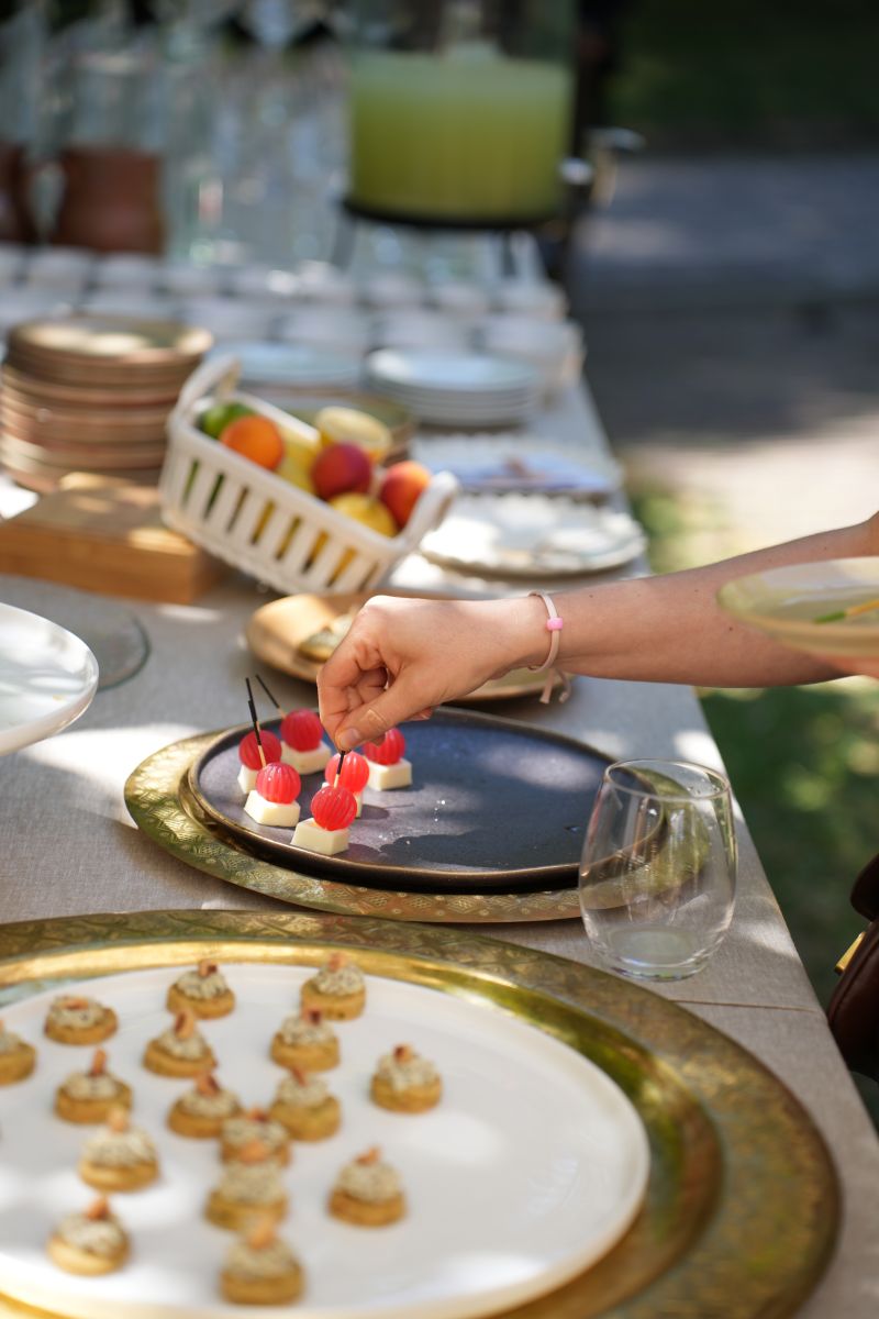 Une main verse une boisson rose rafraîchissante dans un verre sur une table élégante en extérieur, entourée de fruits, de fleurs et de plats colorés, illustrant une ambiance conviviale et estivale.
