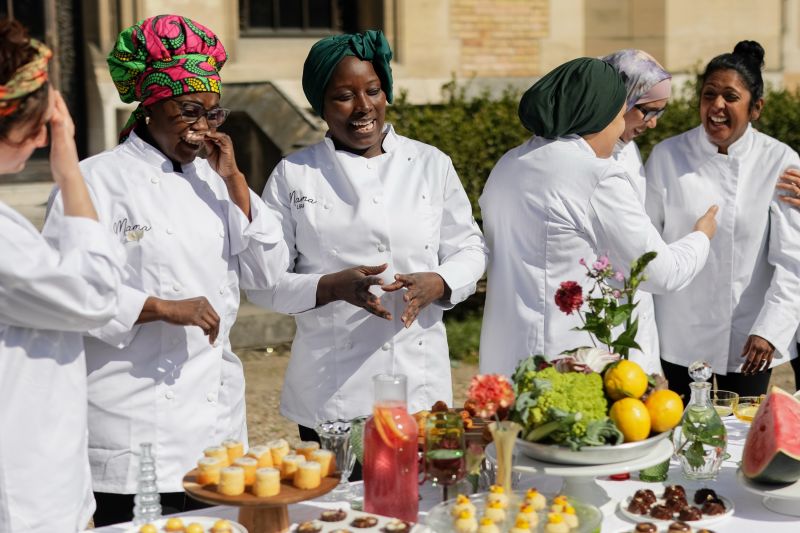 Une main verse une boisson rose rafraîchissante dans un verre sur une table élégante en extérieur, entourée de fruits, de fleurs et de plats colorés, illustrant une ambiance conviviale et estivale.
