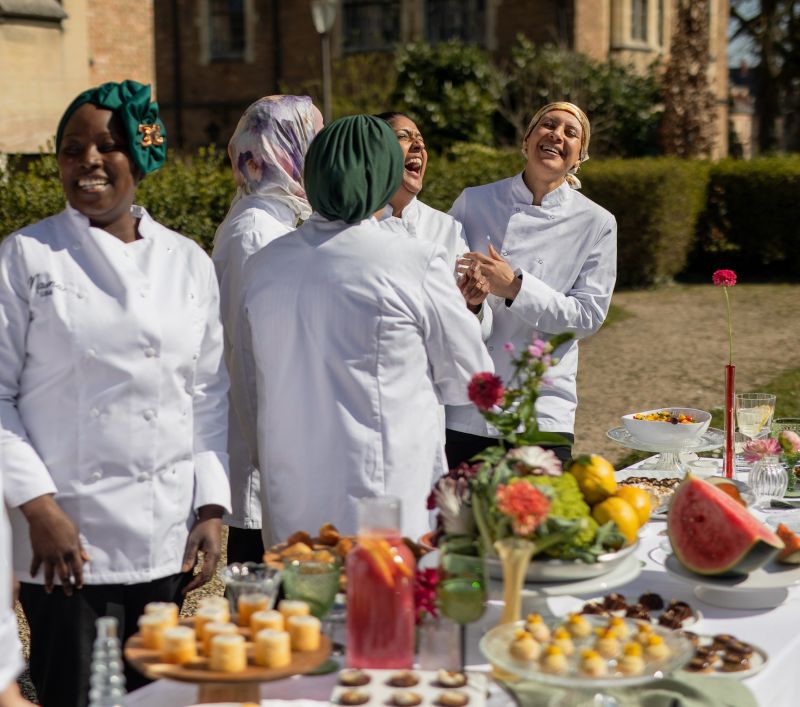 Une cheffe souriante en tenue blanche dresse une table élégante en extérieur avec des amuse-bouches, des plats colorés et des compositions florales, illustrant une expérience traiteur printanière raffinée et conviviale.