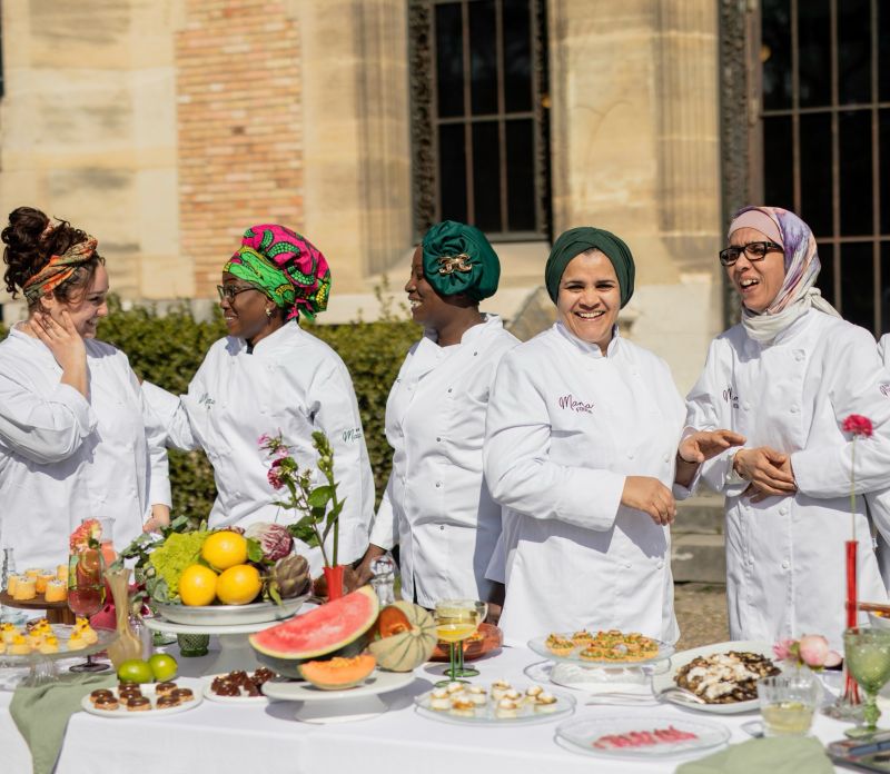 Une table extérieure élégante garnie de fruits frais, légumes de saison, pastèque, agrumes et bouchées apéritives, accompagnée de compositions florales colorées, illustrant une expérience traiteur printanière fraîche et conviviale.