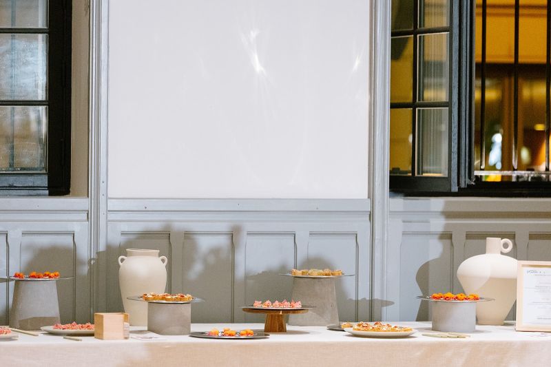 Une table extérieure élégante garnie de fruits frais, légumes de saison, pastèque, agrumes et bouchées apéritives, accompagnée de compositions florales colorées, illustrant une expérience traiteur printanière fraîche et conviviale.